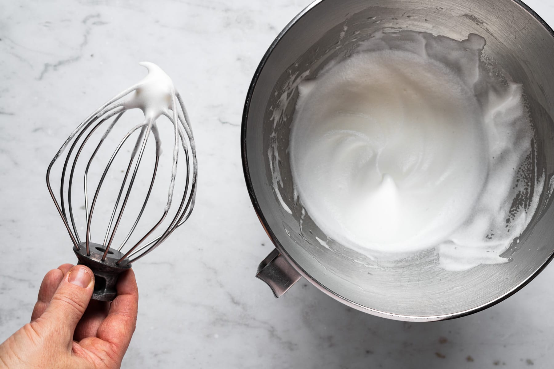 A process photo showing the wire whisk from a stand mixer holding egg whites beaten to soft peaks. The metal bowl of the mixer sits on a marble surface on the right of the photo.
