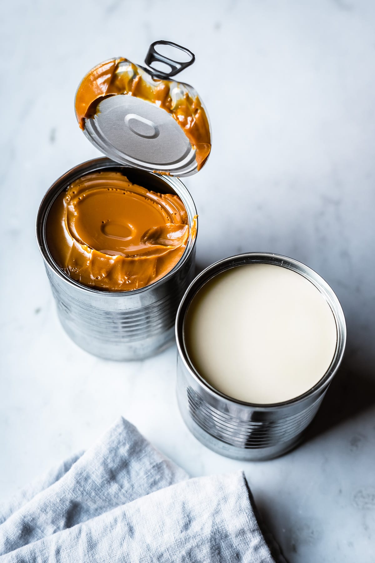 Two open cans, one of condensed milk, the other of dulce de leche, sit on a grey marble surface with a light blue linen napkin nearby.