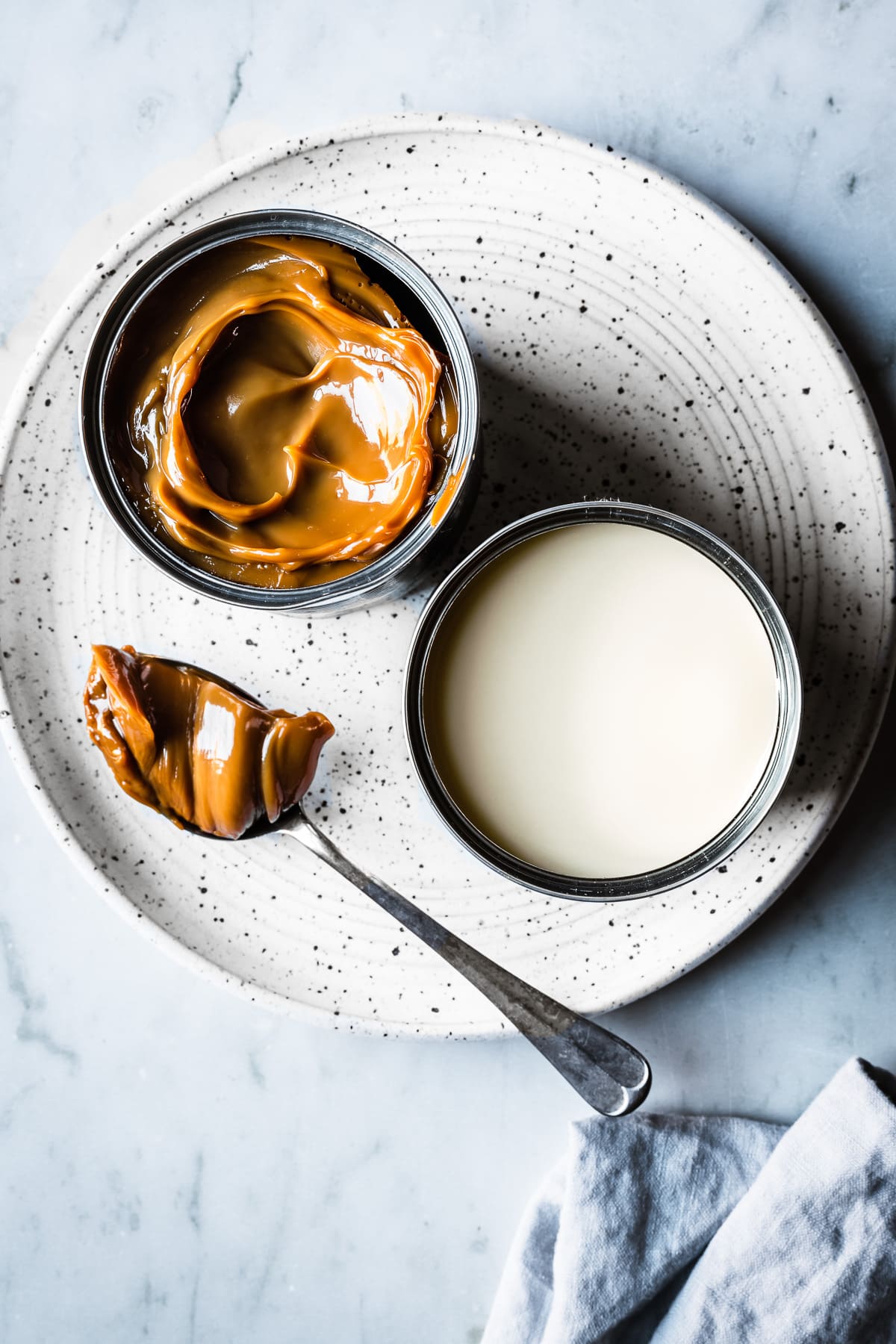 Two open cans, one of condensed milk, the other of dulce de leche, sit on a white speckled ceramic plate on a grey marble surface. A spoon holds a scoop of dulce de leche. A light blue linen napkin peeks into the bottom of the photo.