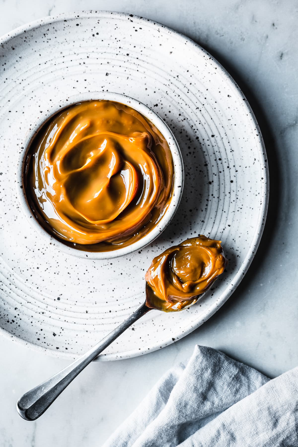 A white ceramic bowl with milk caramel sauce on a white speckled ceramic plate on a grey marble surface. A spoon holds a scoop of dulce de leche. A light blue linen napkin peeks into the bottom of the photo.