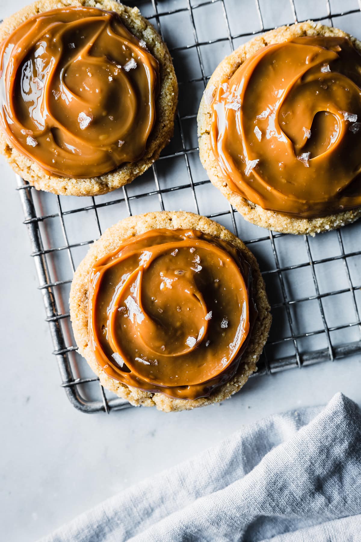 Cookies spread with milk caramel and sprinkled with sea salt on a cooling rack. The rack sits on a grey marble surface. A pale blue linen napkin peeks into the photo at bottom.