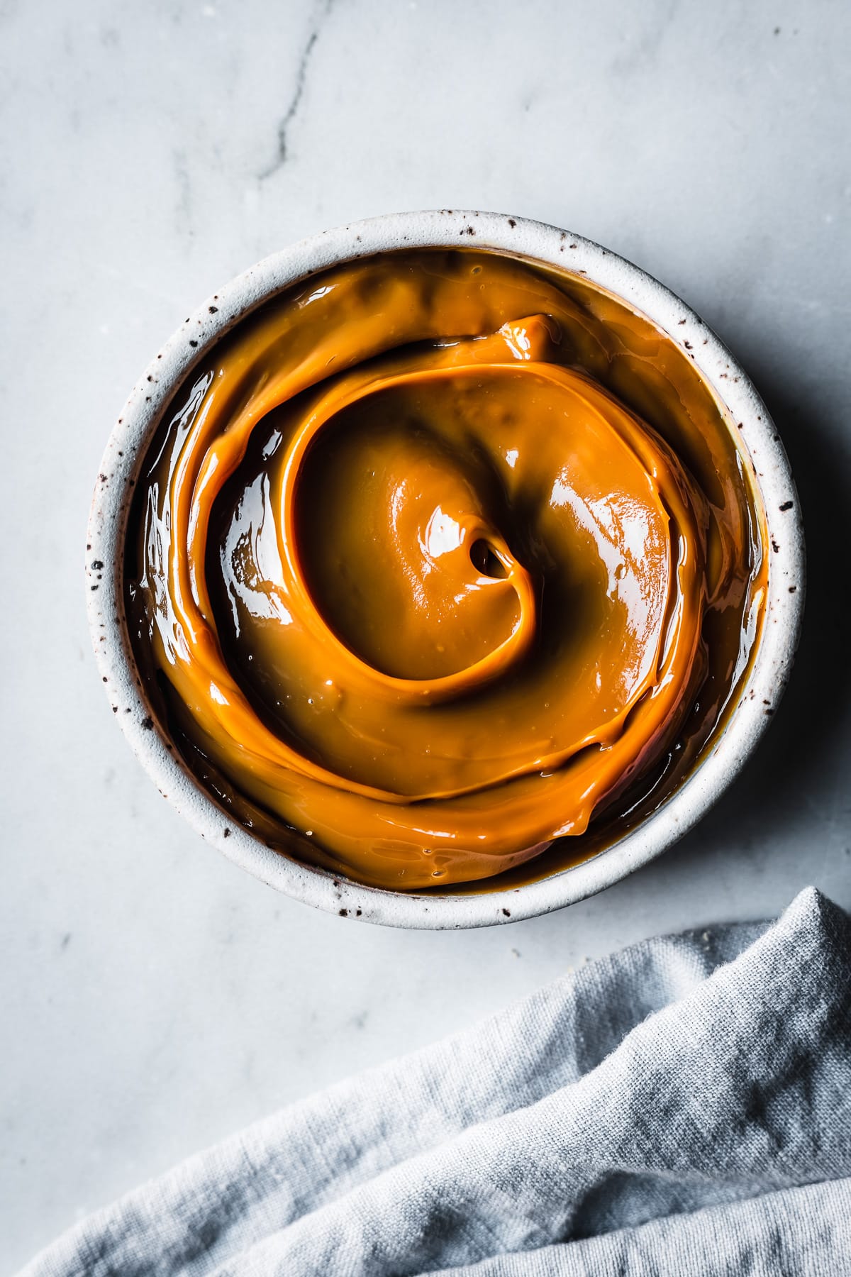 A white ceramic bowl filled with swirls of dulce de leche resting on a grey marble background. A pale blue linen napkin peeks into the bottom of the frame.