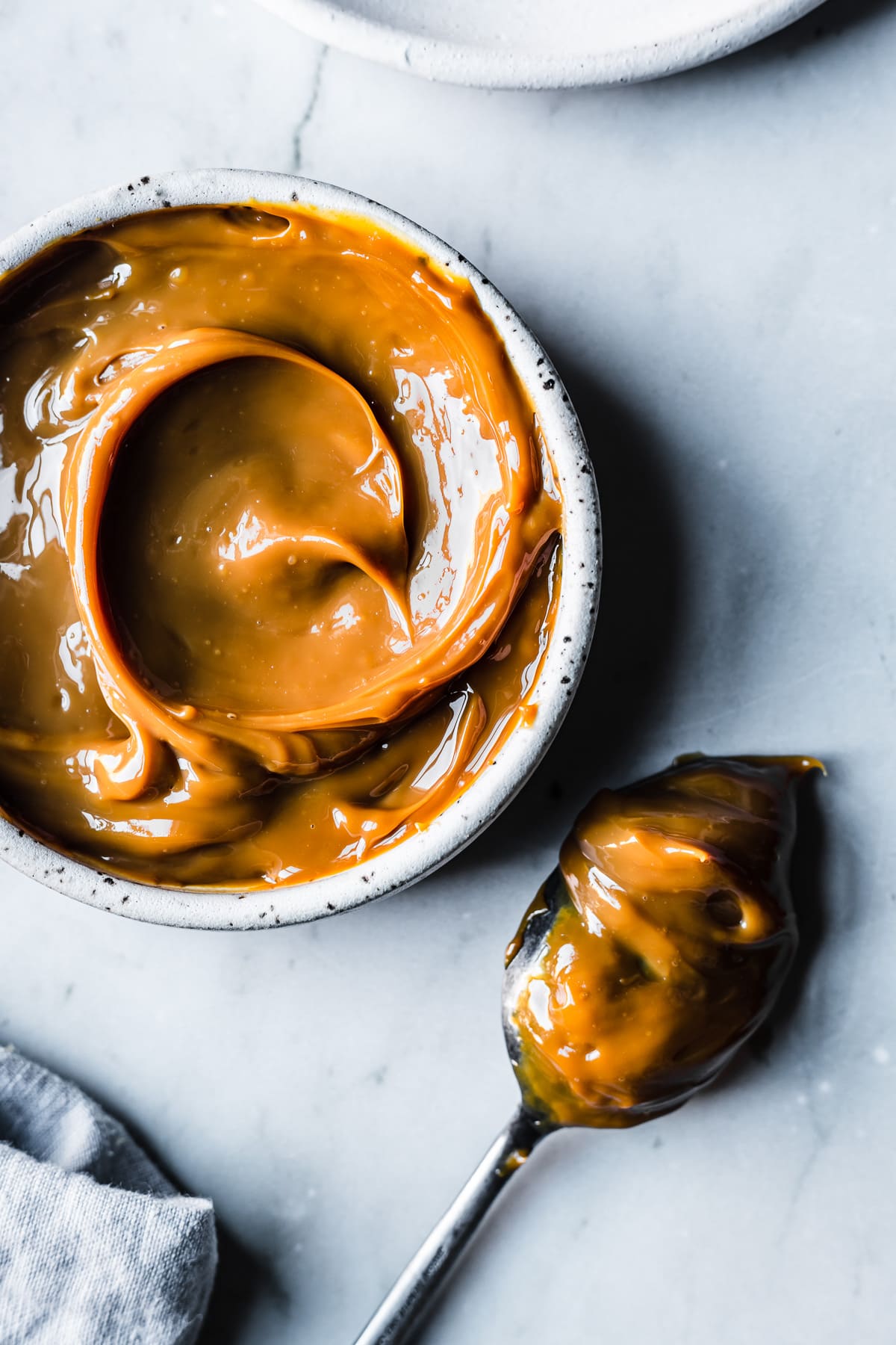 A white ceramic bowl filled with swirls of dulce de leche resting on a grey marble surface. A spoon covered in dulce de leche rests nearby. A pale blue linen napkin peeks into the bottom of the frame.