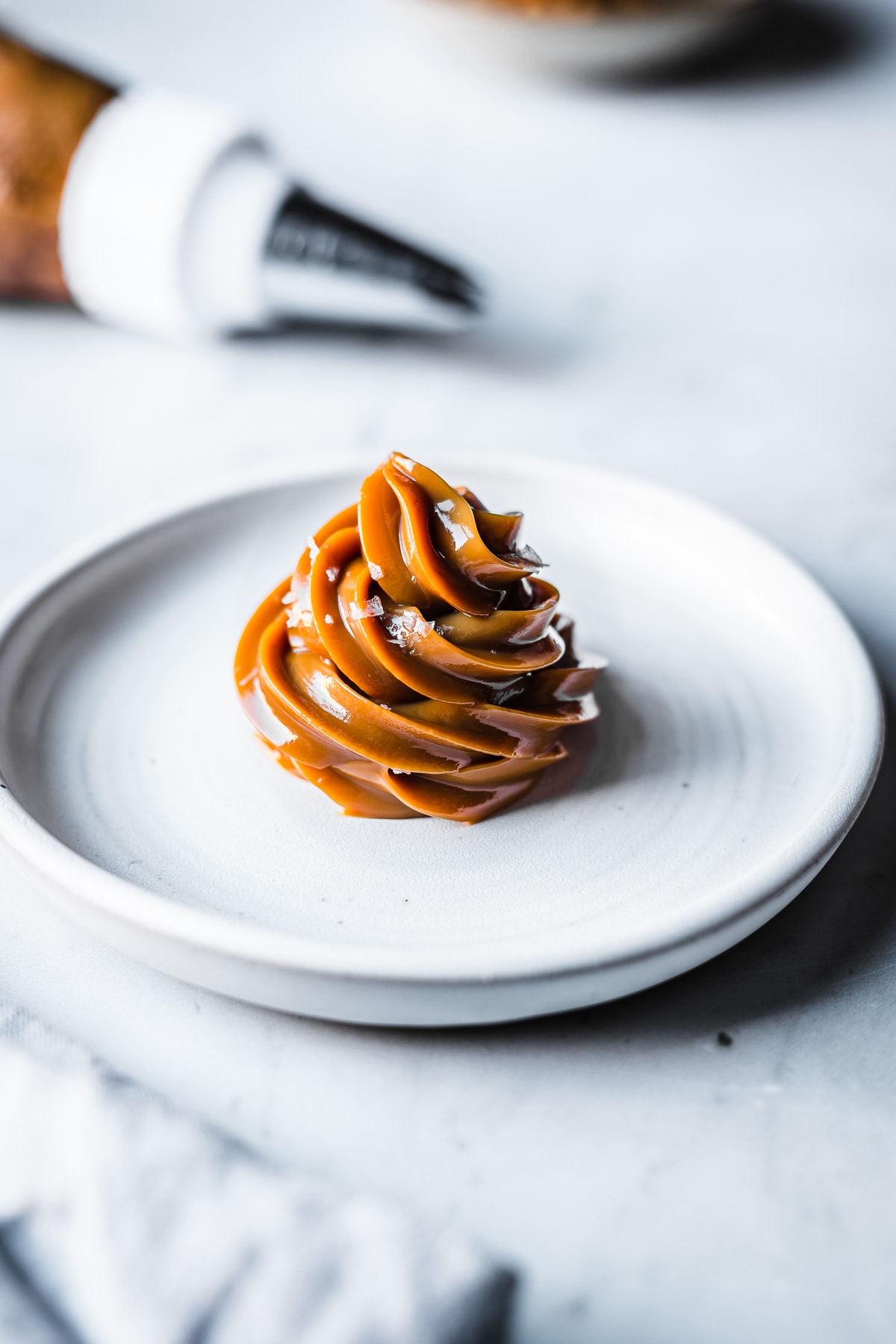 Piped dulce de leche on a white ceramic plate, sprinkled with sea salt flakes. A piping bag rests out of focus in the background. A blurry pale blue napkin peeks into the foreground.