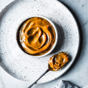 A white ceramic bowl with milk caramel sauce on a white speckled ceramic plate on a grey marble surface. A spoon holds a scoop of dulce de leche. A light blue linen napkin peeks into the bottom of the photo.