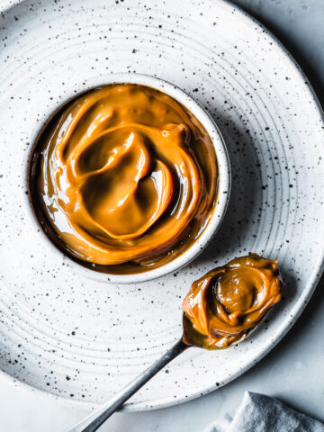 A white ceramic bowl with milk caramel sauce on a white speckled ceramic plate on a grey marble surface. A spoon holds a scoop of dulce de leche. A light blue linen napkin peeks into the bottom of the photo.