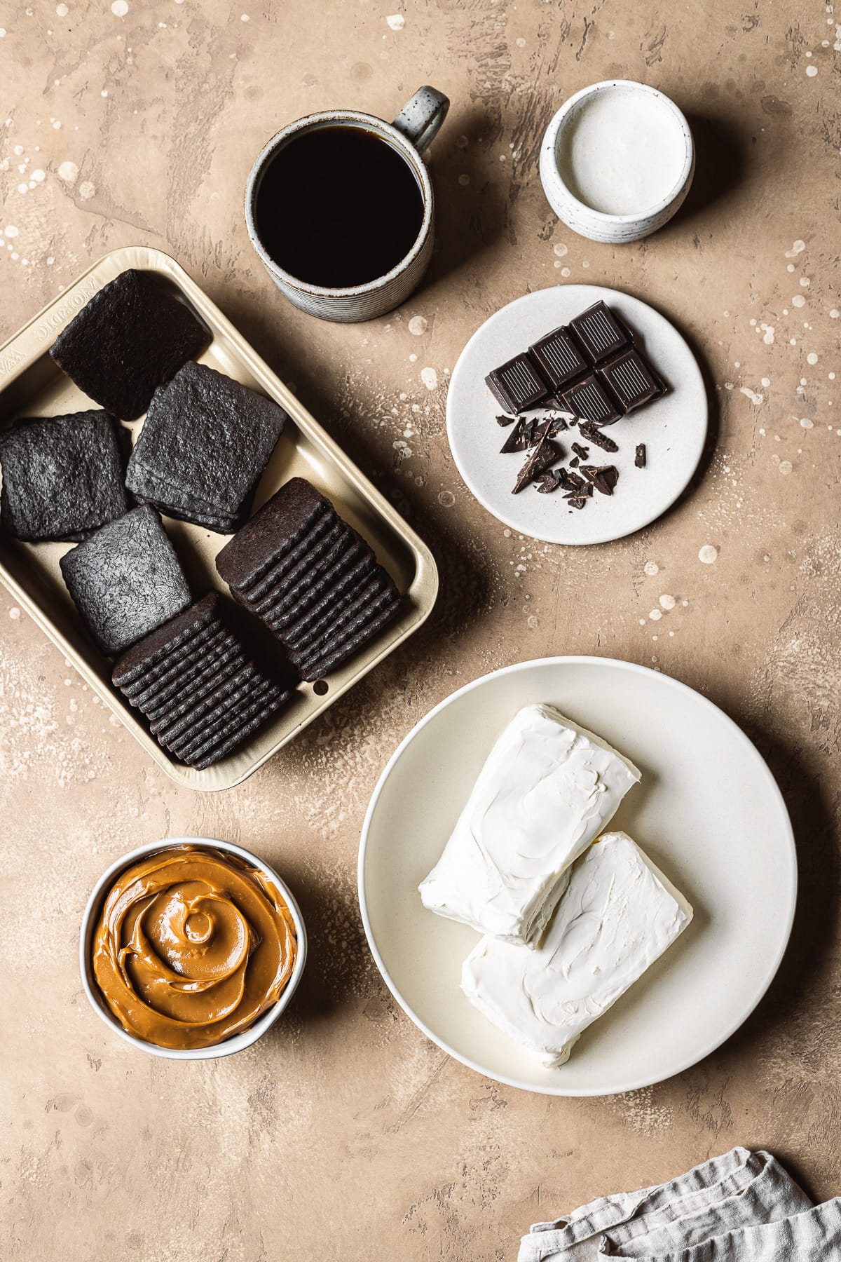 A photo of the ingredients required to make an Argentine chocotorta, including cream cheese, chocolate wafer cookies and dulce de leche. The ingredients are in individual containers on a textured tan stone surface.