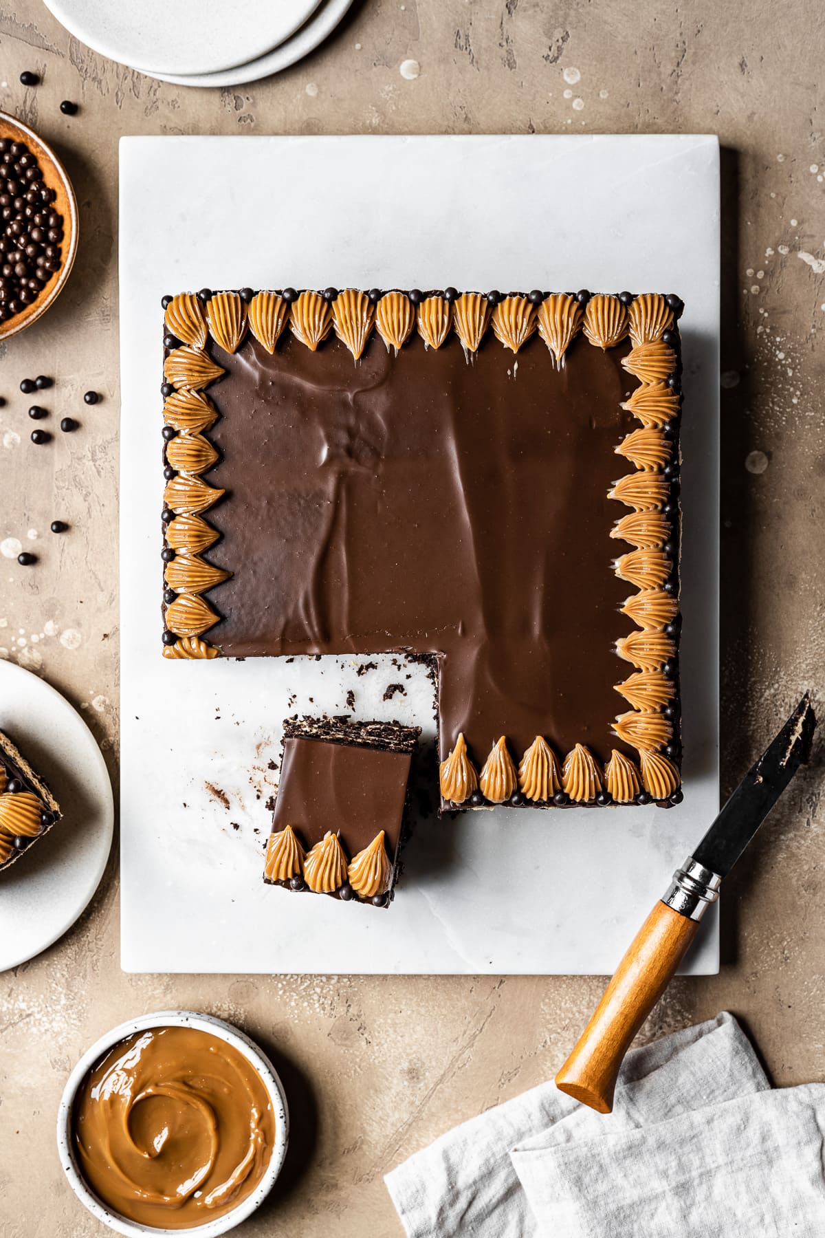 A square chocolate icebox cake has two slices cut out of it. The chocotorta has a top layer of chocolate ganache and piped dulce de leche decorations on a rectangular white marble platter. Small plates, bowls of ingredients and a linen napkin and knife rest nearby partially out of frame.