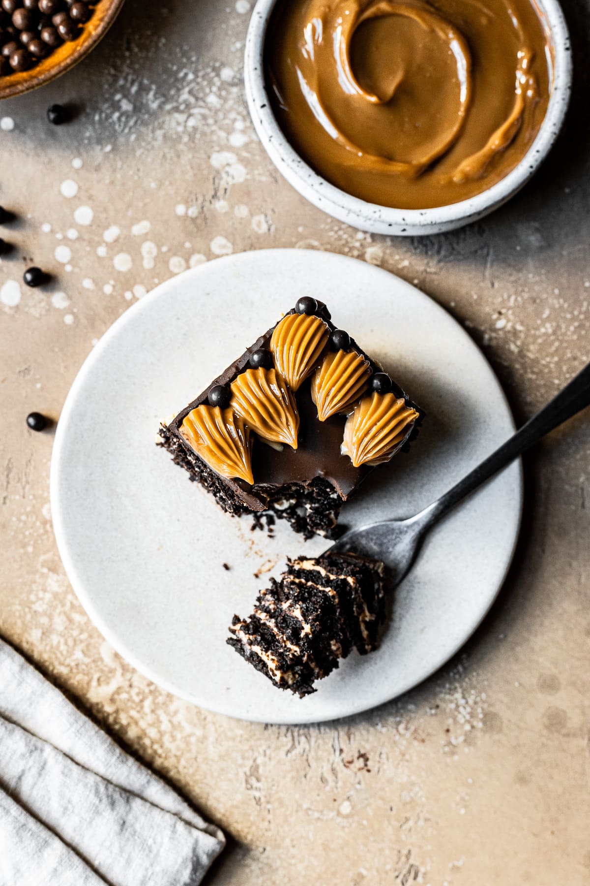 A square piece of chocolate dulce de leche icebox cake on a beige ceramic plate on a tan stone surface. There is a bit of cake resting on a fork on the plate. A small white ceramic bowl of dulce de leche is partially out of the frame at top right.