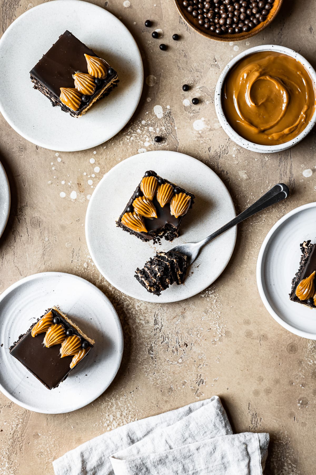 Square slices of chocolate dulce de leche icebox cake on white ceramic plates on a tan stone surface. One slice has a bite of cake resting on a fork. Bowls of chocolate pearls and dulce de leche are at top left. A tan linen napkin rests at bottom.