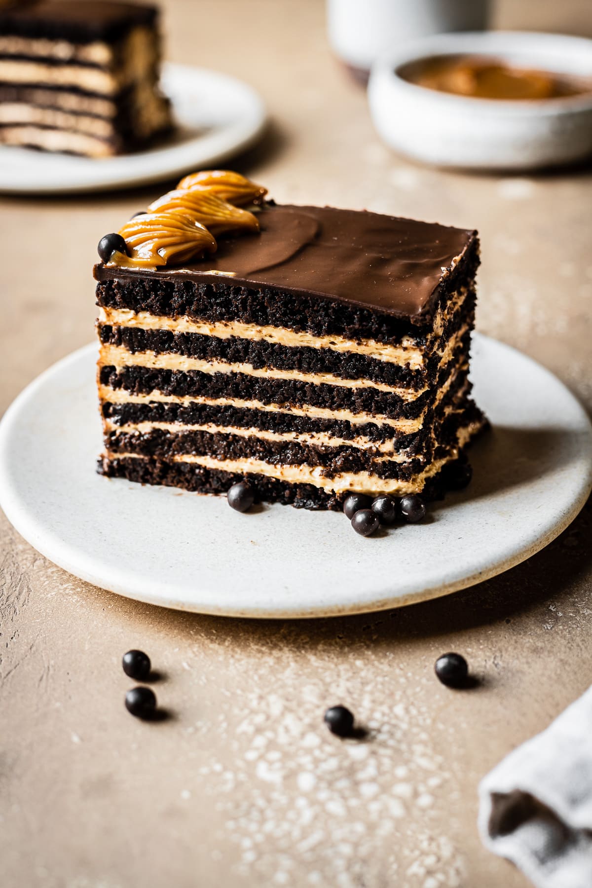 A side view of a rectangular piece of chocotorta icebox cake showing the alternating chocolate cookie layers and the dulce de leche cream cheese layers. The cake rests on a beige ceramic plate on a tan stone surface. Another slice and a bowl of dulce de leche are out of focus in the background.