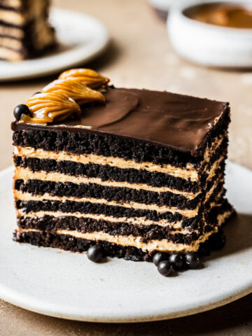A side view of a rectangular piece of chocotorta icebox cake showing the alternating chocolate cookie layers and the dulce de leche cream cheese layers. The cake rests on a beige ceramic plate on a tan stone surface. Another slice and a bowl of dulce de leche are out of focus in the background.