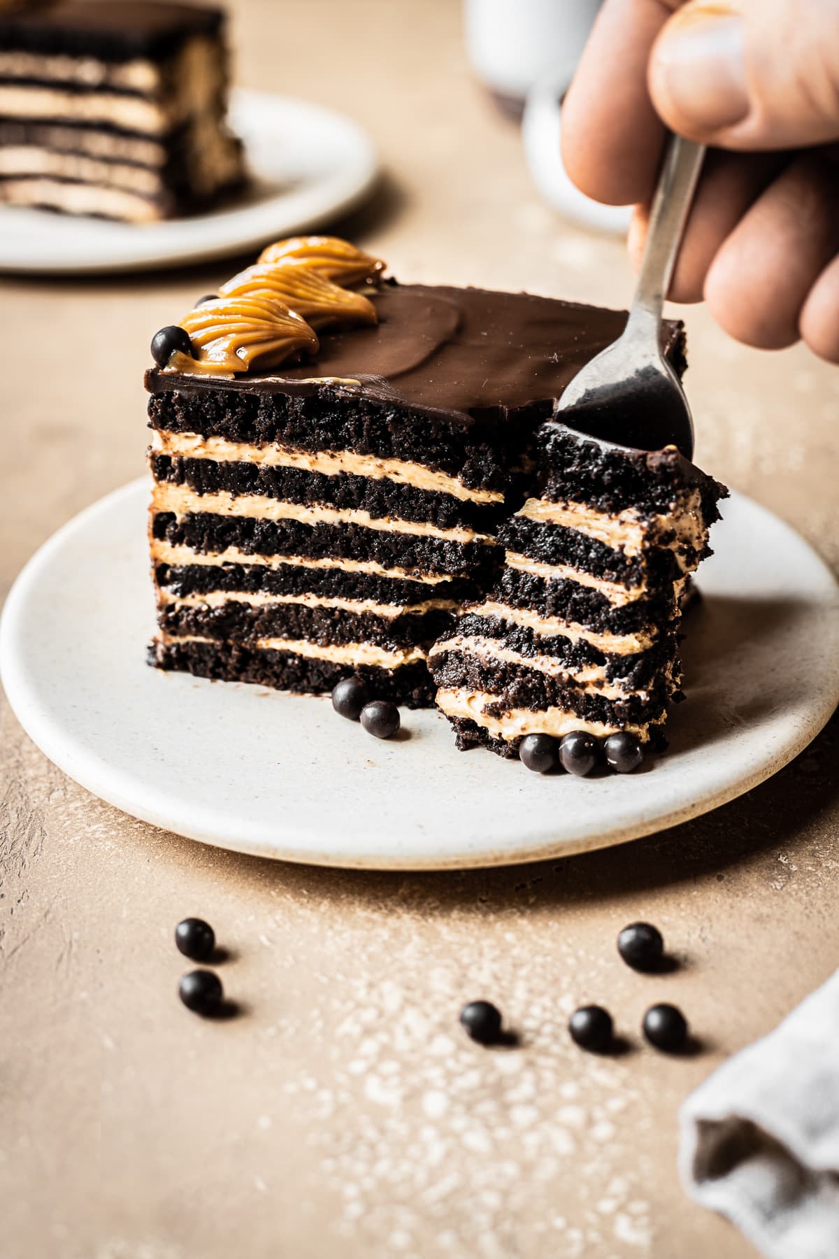 A side view of a rectangular piece of chocotorta icebox cake showing the alternating chocolate cookie layers and the dulce de leche cream cheese layers. The cake rests on a beige ceramic plate on a tan stone surface. A hand reaches into the frame, holding a fork that is scooping a bite of cake onto it. Another slice and a bowl of dulce de leche are out of focus in the background.