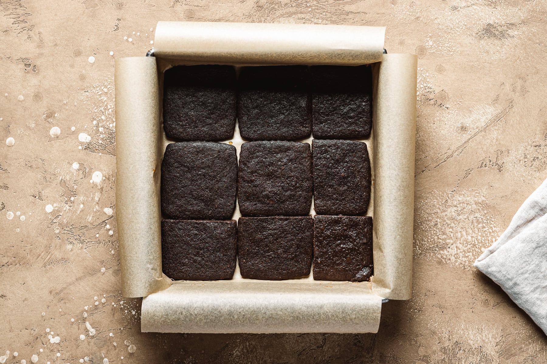 A recipe process photo showing a layer of chocolate wafer cookies in a square pan. The pan sits on a tan textured stone surface.
