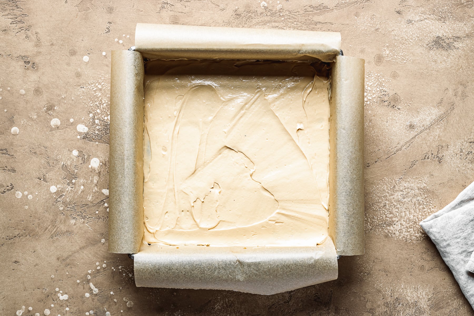 A recipe process photo showing a smooth layer of dulce de leche filling in a square pan. The pan sits on a tan textured stone surface.