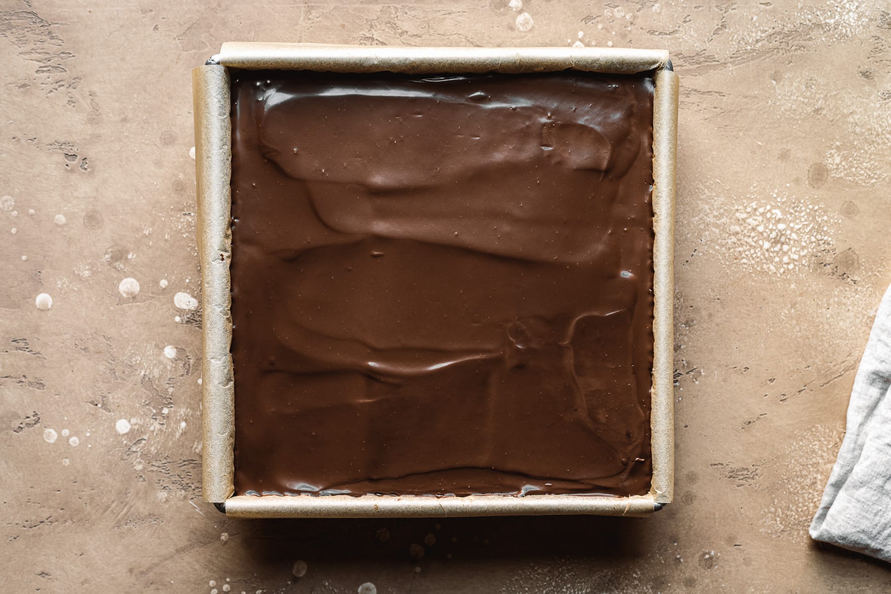 A recipe process photo showing a top layer of dark chocolate ganache in a square pan. The pan sits on a tan textured stone surface.