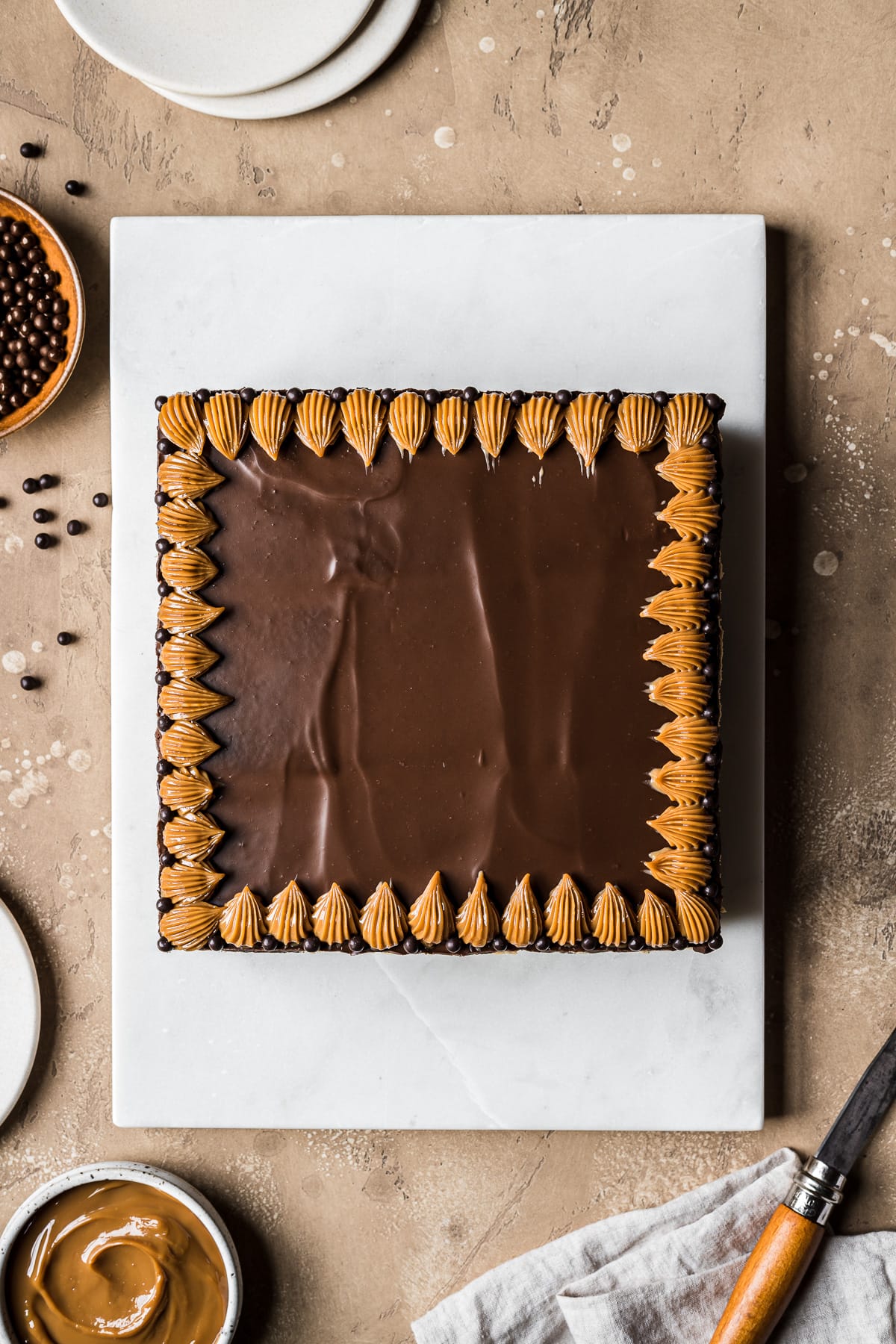 A square chocolate icebox cake with a top layer of chocolate ganache and piped dulce de leche decorations on a rectangular white marble platter. Small plates, bowls of ingredients and a linen napkin and knife rest nearby partially out of frame.