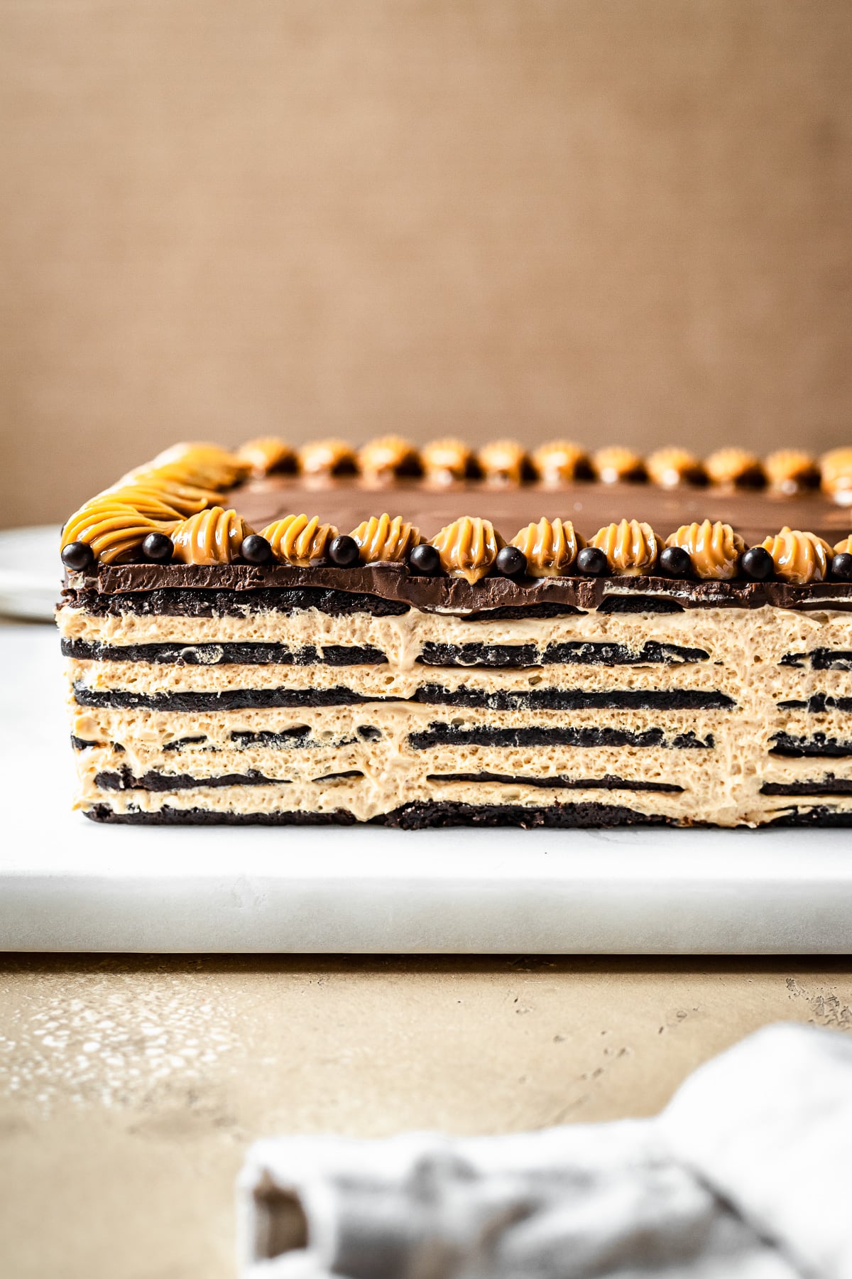 A side view of a chocotorta revealing layers of chocolate wafer cookies alternating with dulce de leche cream cheese filling. A layer of chocolate ganache tops the cake, with decorative dulce de leche piping and chocolate pearls. The cake rests on a white marble board on a tan stone surface. The background is a warm tan color.