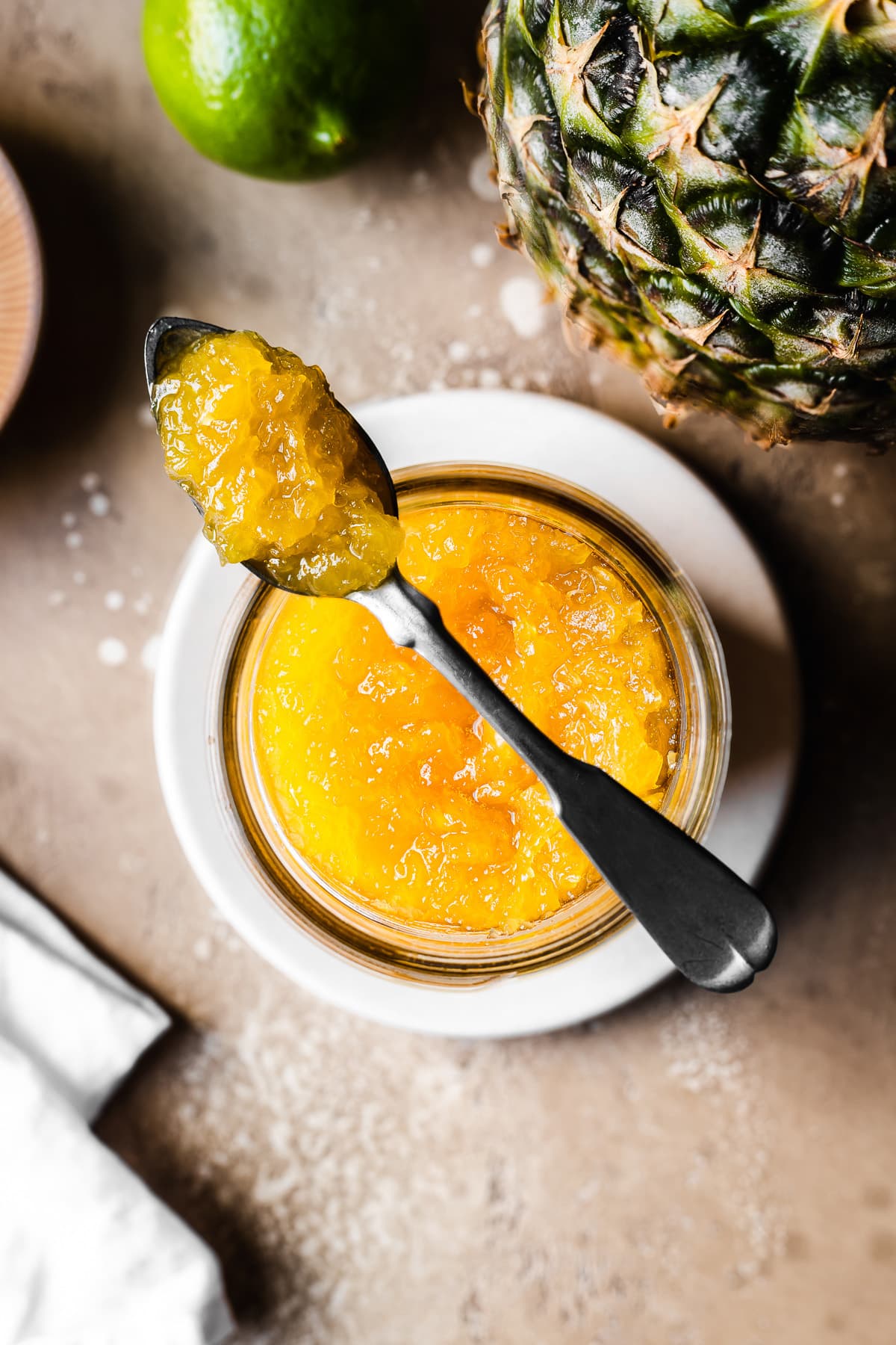 A spoonful of pineapple jam rests atop a jar of jam on a small plate. There is a pineapple peeking into the photo at top right along with a lime. The surface is tan stone.