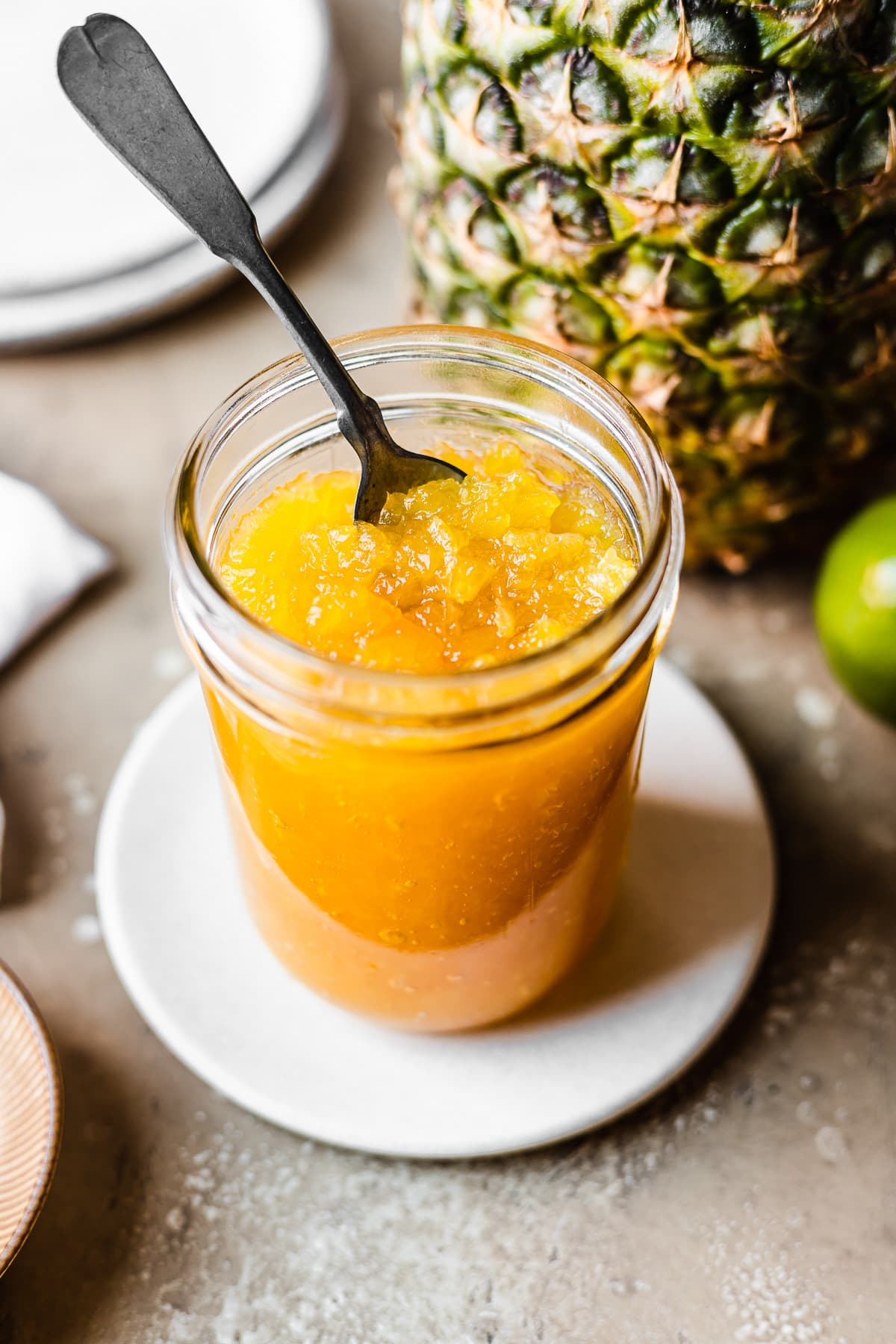 45 degree angle view of a glass jar of pineapple jam with a vintage silver spoon inside. The jar sits on a small tan plate on a warm stone surface. It is surrounded by a pineapple, lime, plates and a white linen napkin.