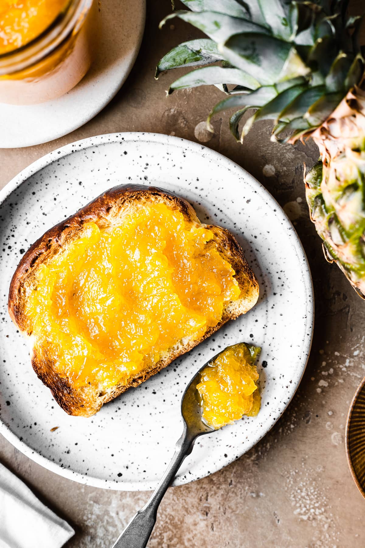 A piece of toast spread with bright yellow pineapple jam on a white speckled ceramic plate. There is a spoonful of jam resting on the plate. A pineapple and a glass mason jar of jam peek into the photo at top.