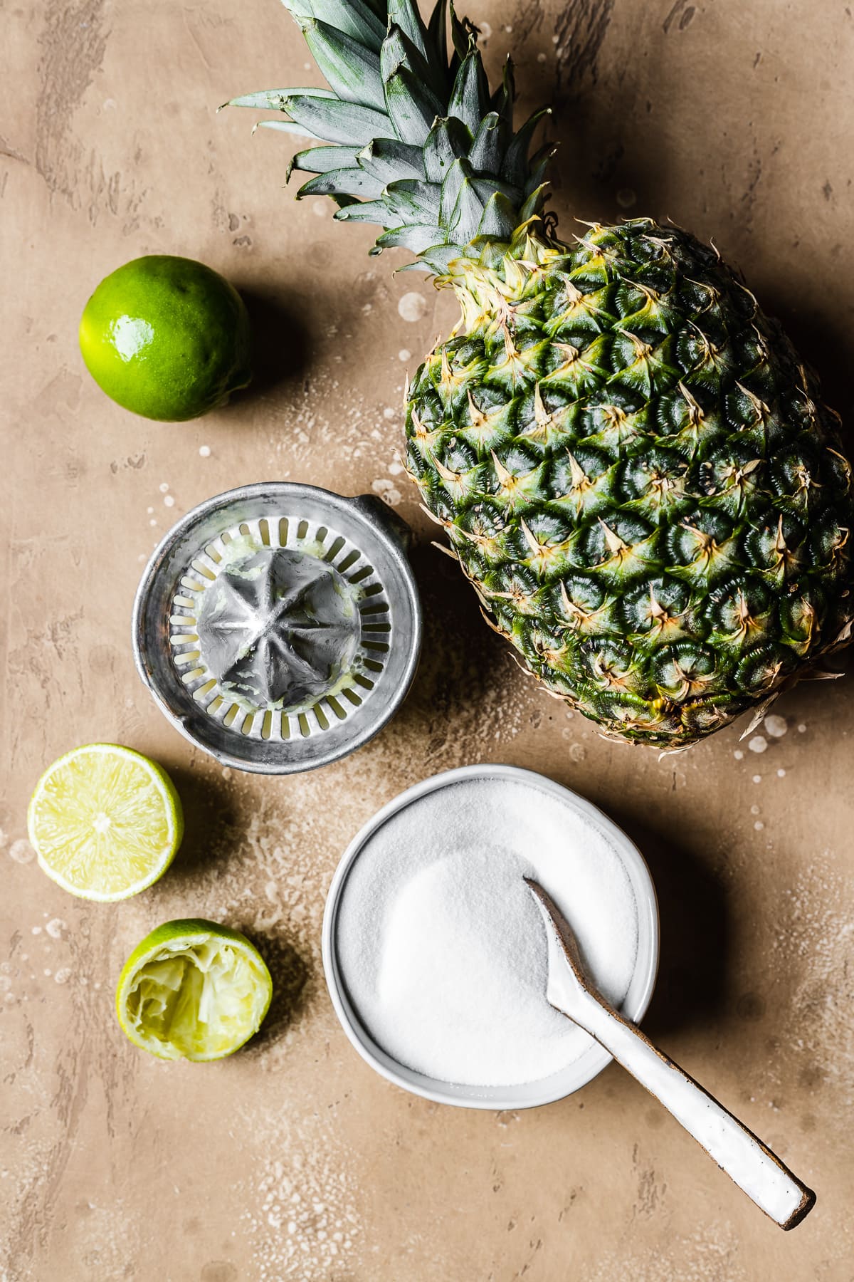 A photo showing the ingredients for pineapple jam: a fresh pinapple, granulated sugar in a white ceramic bowl with a white spoon, and a vintage metal juicer with a whole lime and a squeezed lime nearby. The items rest on a speckled tan stone surface.