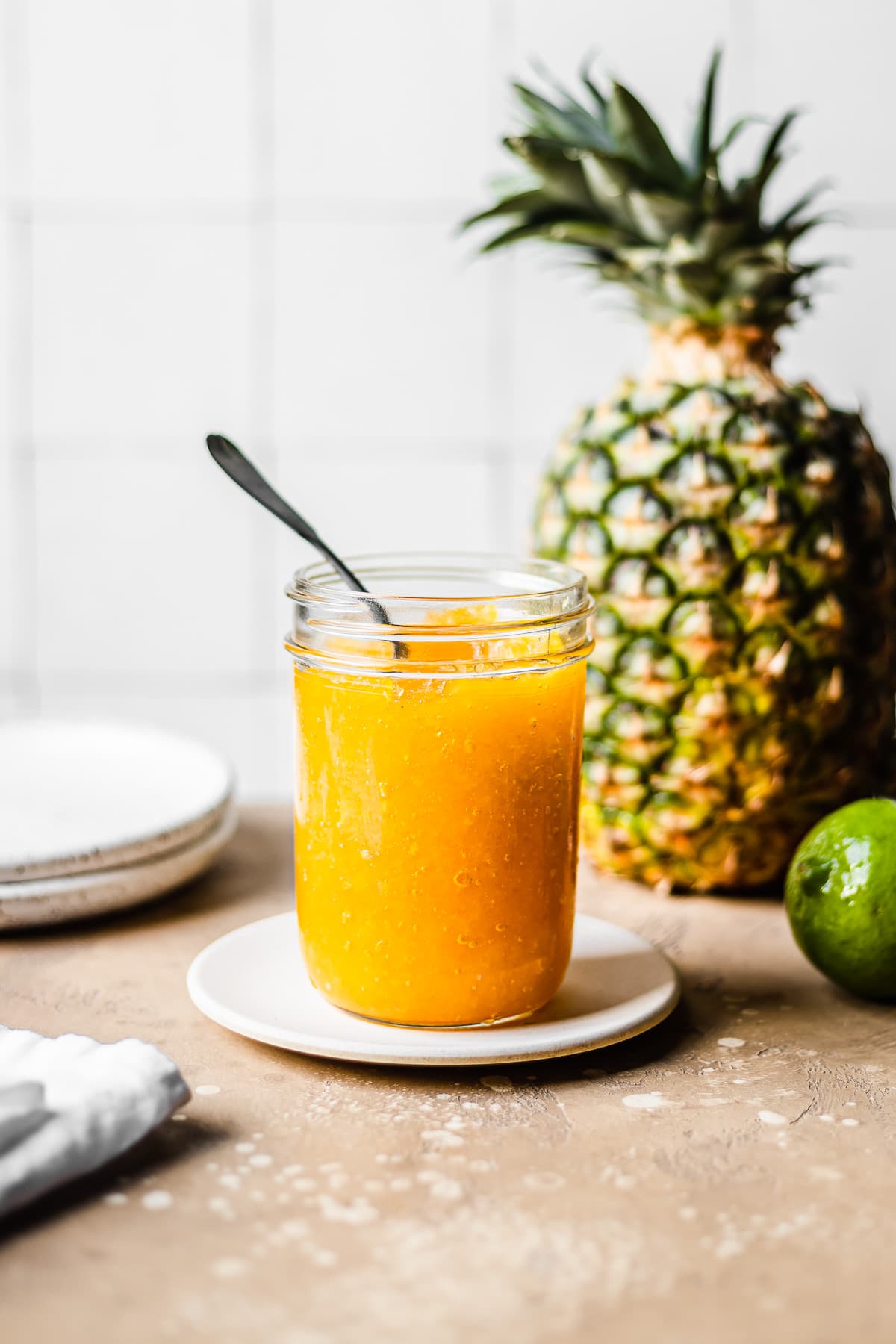 A mason jar of golden yellow jam sits on a small tan plate on a speckled tan stone surface. In the background are a lime and a pineapple and two plates. A white tile wall is out of focus in the background.