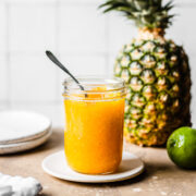 A mason jar of golden yellow jam sits on a small tan plate on a speckled tan stone surface. In the background are a lime and a pineapple and two plates. A white tile wall is out of focus in the background.