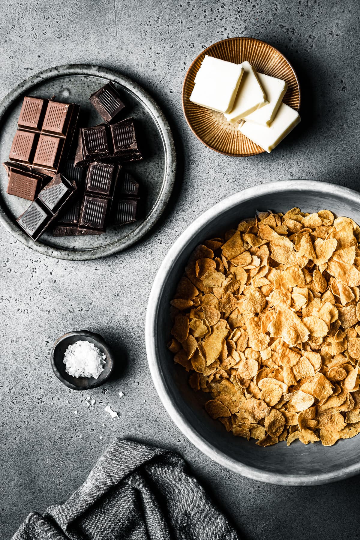 A process photo showing the ingredients for the roses de sables recipe, including a bowl of cornflakes, cubed butter, sea salt and two types of chocolate. The containers rest on a grey stone surface with a grey napkin nearby.