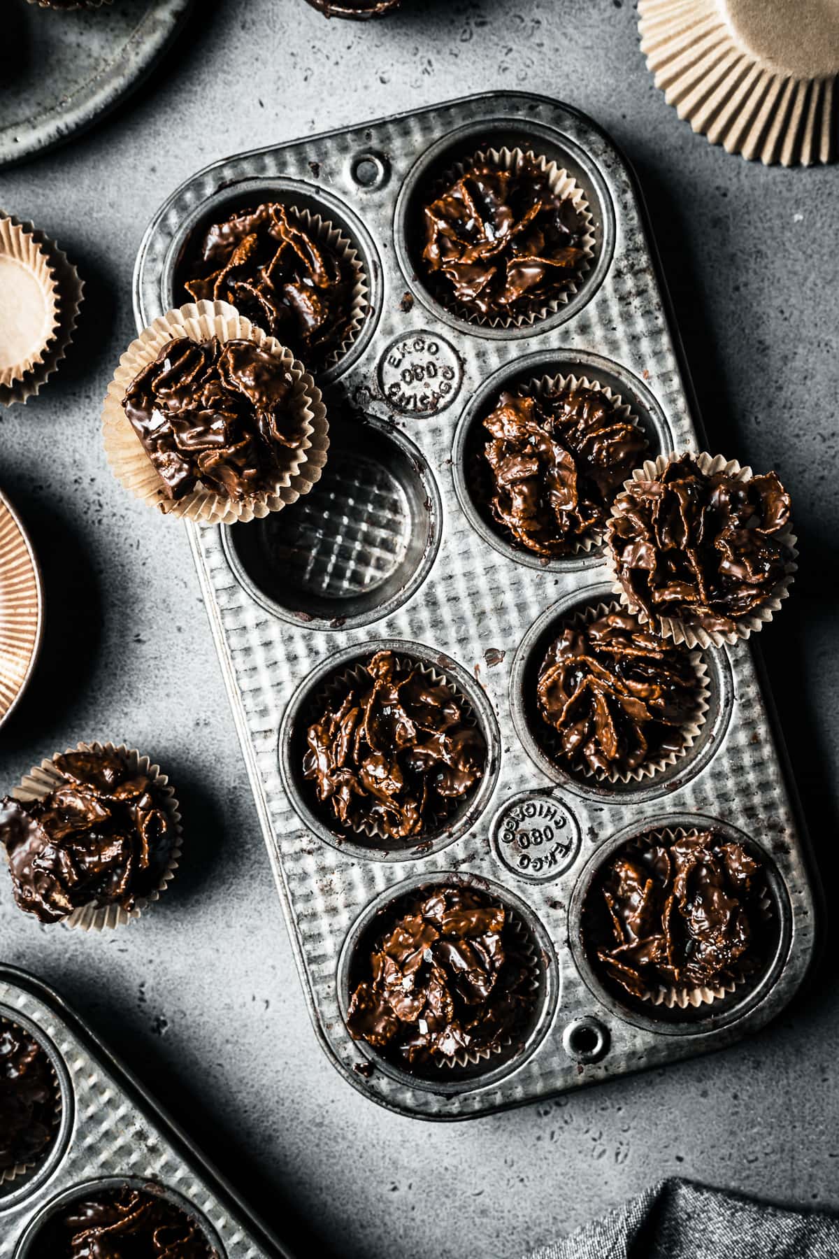A vintage silver mini muffin tins hold chocolate covered cornflake cereal, called roses de sables. They rest on a grey stone surface and are surrounded by a brown bowl and brown paper muffin liners,