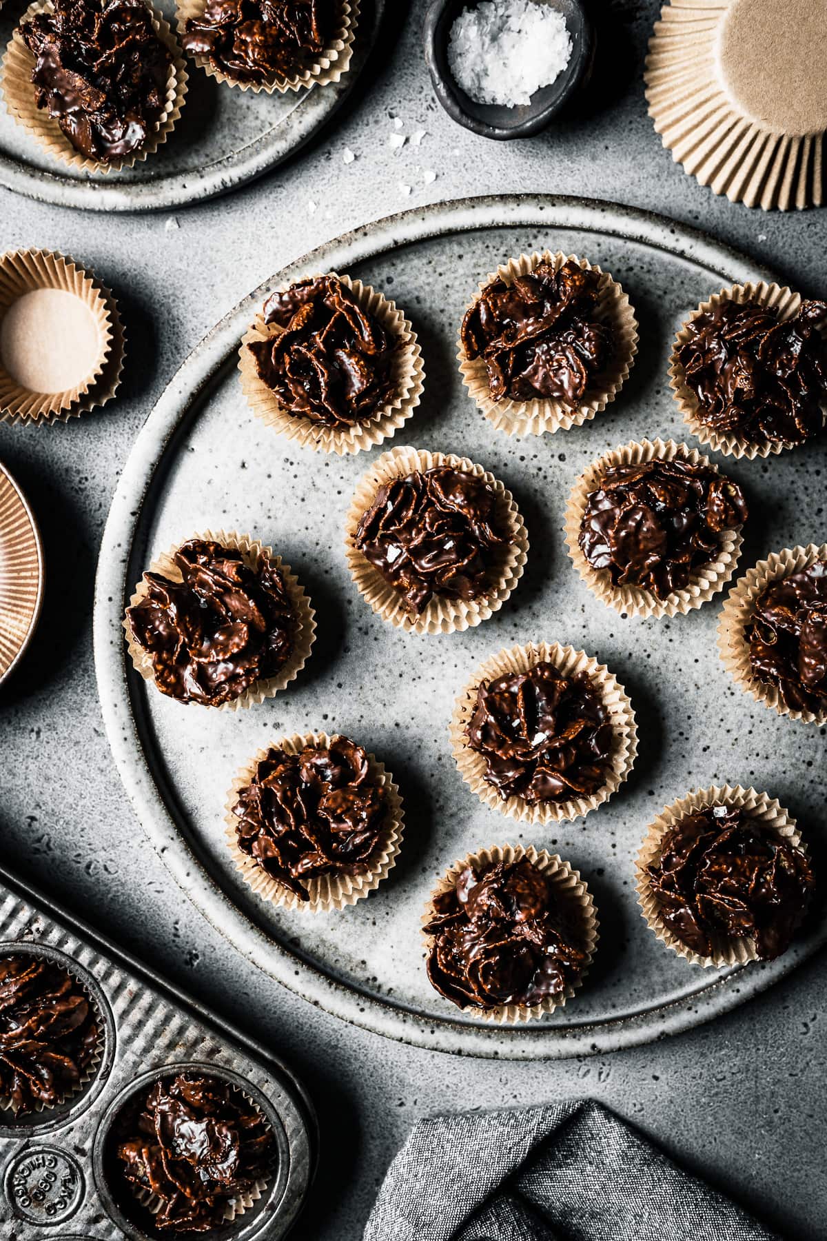 A large speckled grey ceramic plate holds chocolate covered cornflake cereal in doubled up mini brown paper cupcake liners. They rest on a grey stone surface and are surrounded by another muffin tin, a brown bowl and brown paper cupcake liners,