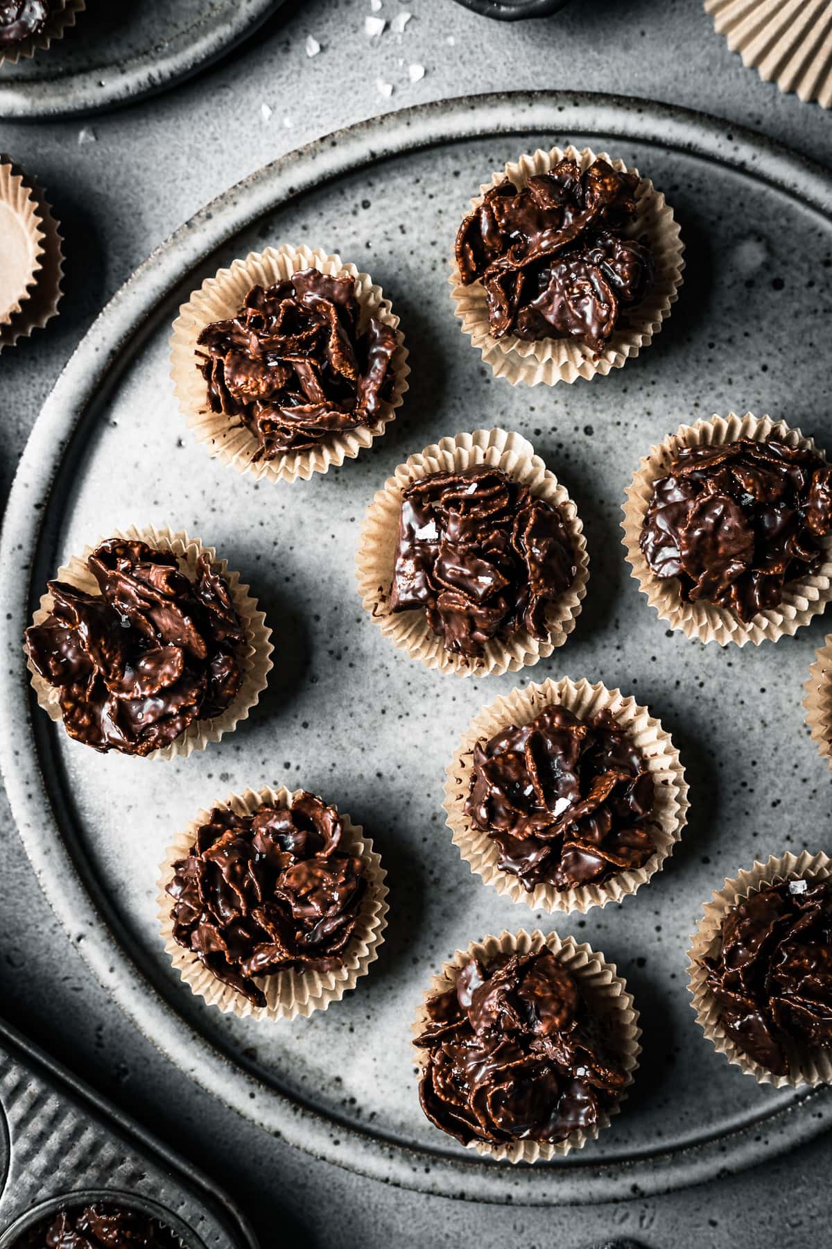 A large speckled grey ceramic plate holds chocolate covered cornflake cereal in doubled up mini brown paper cupcake liners. They rest on a grey stone surface and are surrounded by another muffin tin, a brown bowl and brown paper cupcake liners,