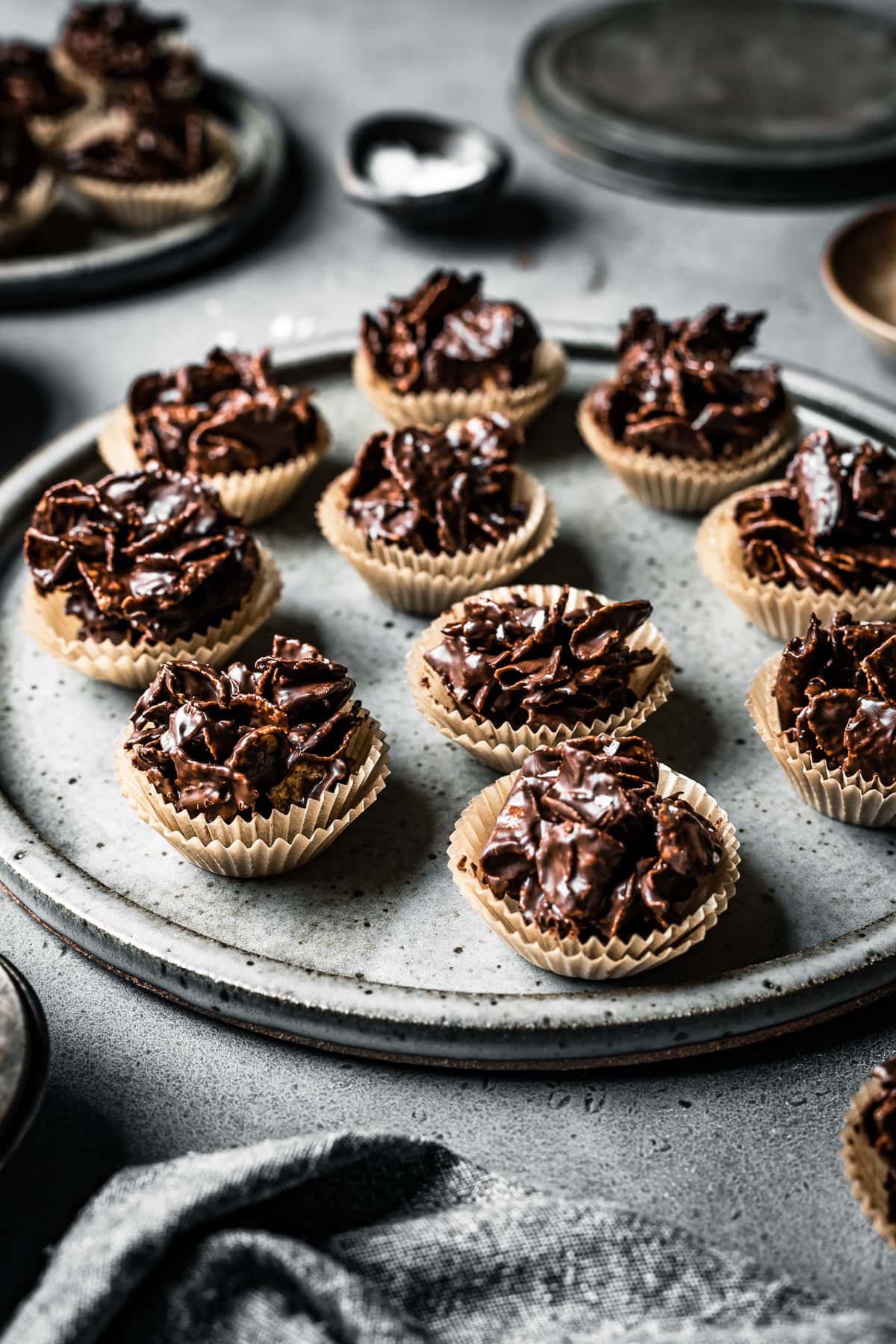 A slde view of a large speckled grey ceramic plate holds chocolate covered cornflake cereal in doubled up mini brown paper cupcake liners. They rest on a grey stone surface and are surrounded bya grey linen napkin, a pinch bowl of flaky salt and another plate of candies.