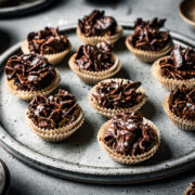 A slde view of a large speckled grey ceramic plate holding chocolate covered cornflake cereal in doubled up mini brown paper cupcake liners. The plate sits on a grey stone surface.