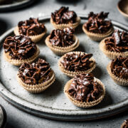 A slde view of a large speckled grey ceramic plate holding chocolate covered cornflake cereal in doubled up mini brown paper cupcake liners. The plate sits on a grey stone surface.