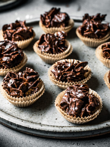 A slde view of a large speckled grey ceramic plate holding chocolate covered cornflake cereal in doubled up mini brown paper cupcake liners. The plate sits on a grey stone surface.