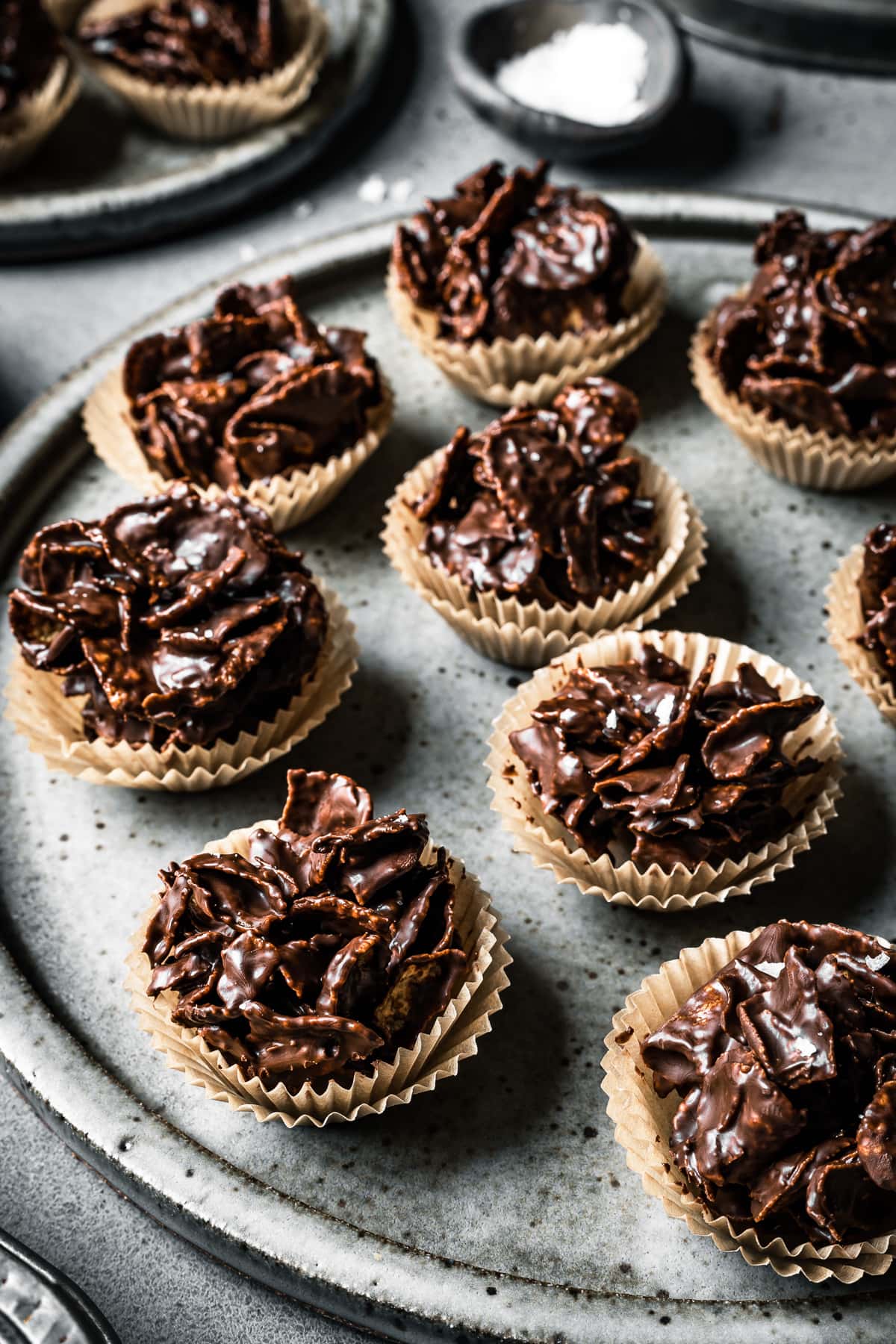 A slde view of a large speckled grey ceramic plate holding chocolate covered cornflake cereal in doubled up mini brown paper cupcake liners. The plate rests on a grey stone surface.