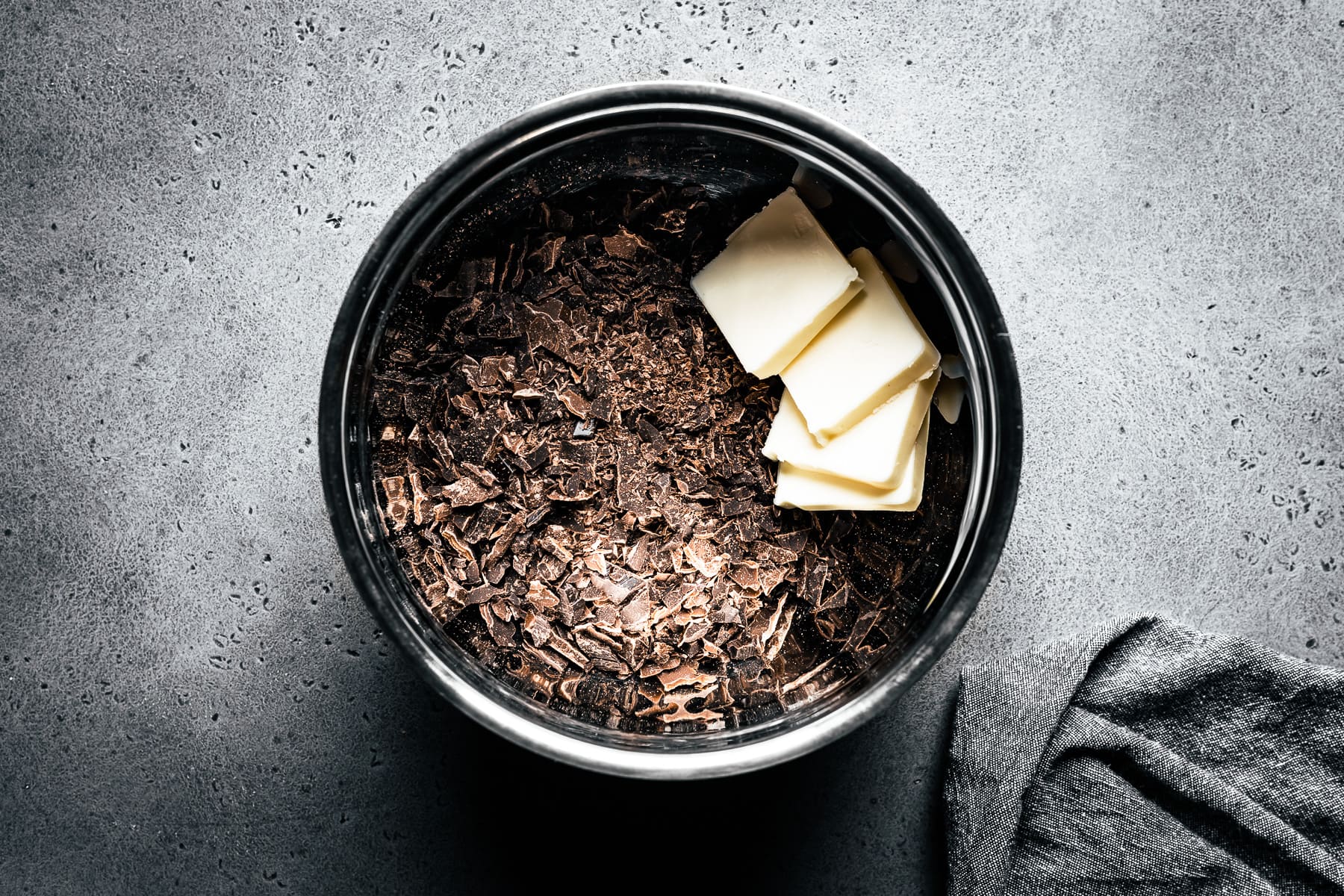A process photo showing a metal bowl with finely chopped chocolate and pats of butter inside. The bowl rests on a grey stone surface with a grey napkin nearby.