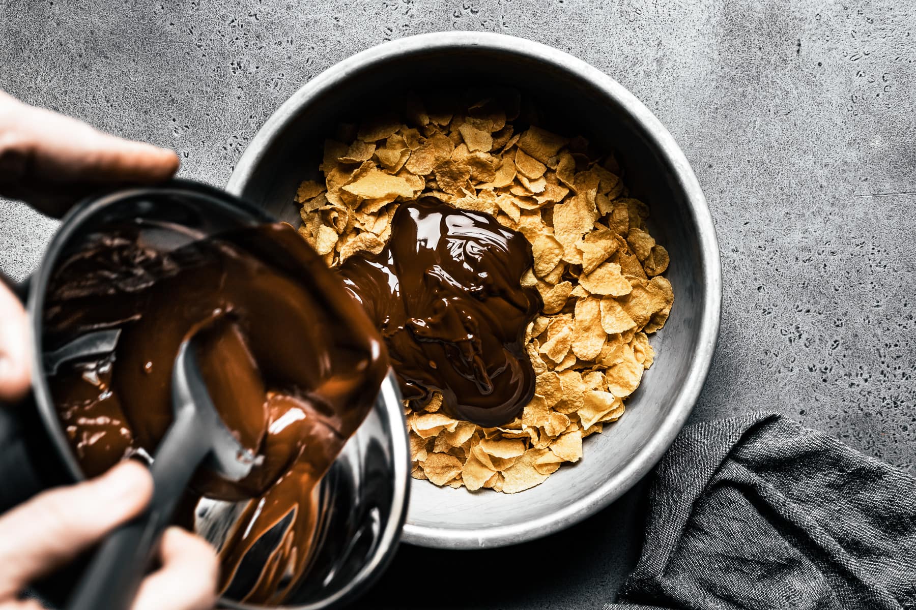 A process photo showing a bowl of melted chocolate being poured onto a larger bowl of cornflakes. The bowl of cereal rests on a grey stone surface with a grey napkin nearby.