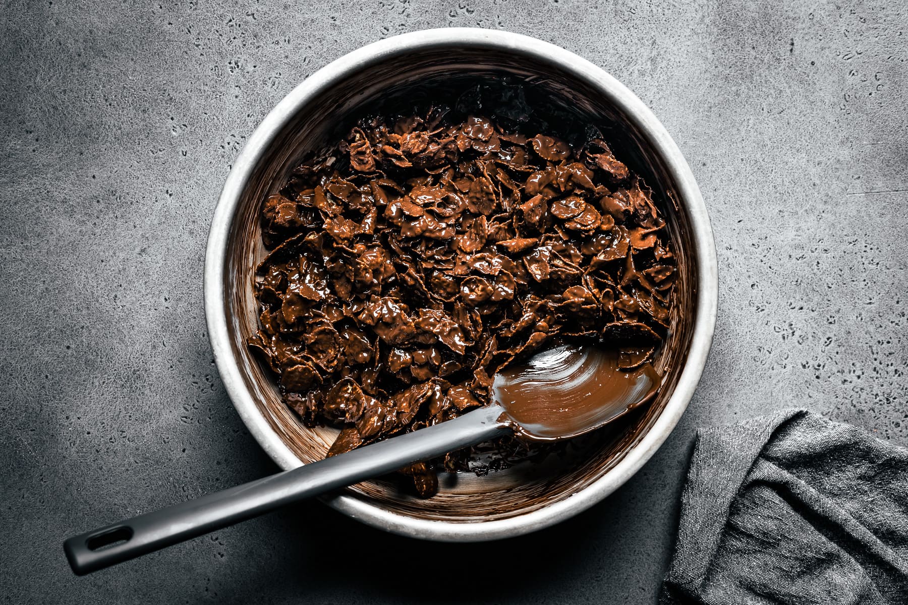 A process photo showing a metal bowl of cereal covered in melted chocolate with a grey spatula resting in it. The bowl rests on a grey stone surface with a grey napkin nearby.