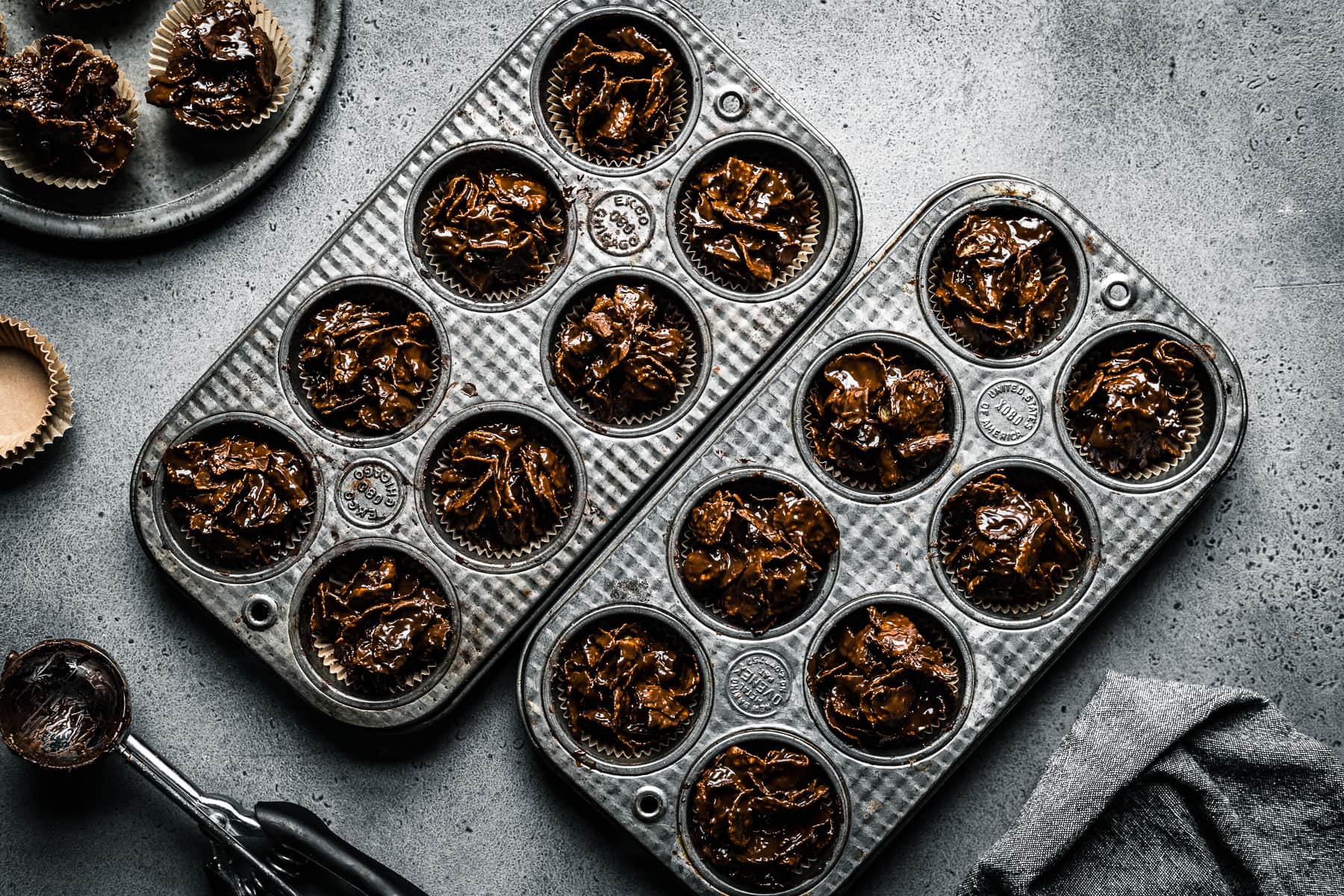 Two vintage mini muffin tins hold chocolate covered cornflake cereal, called roses de sables. They rest on a grey stone surface and are surrounded by a grey ceramic plate, brown paper muffin liners, a cookie scoop and a grey linen napkin.