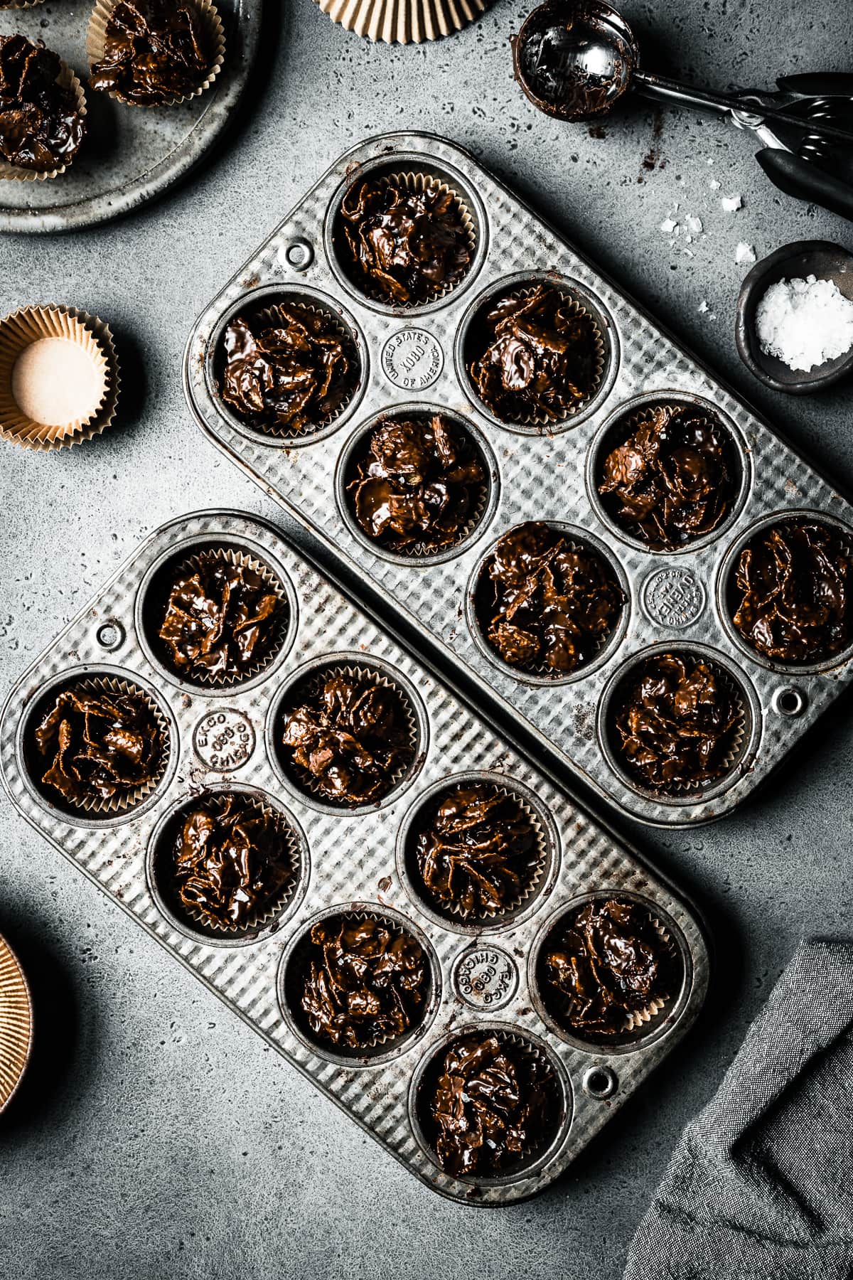 Two vintage mini muffin tins hold chocolate covered cornflake cereal, called roses de sables. They rest on a grey stone surface and are surrounded by brown bowls, brown paper muffin liners, a cookie scoop and a pinch bowl of flaky sea salt.