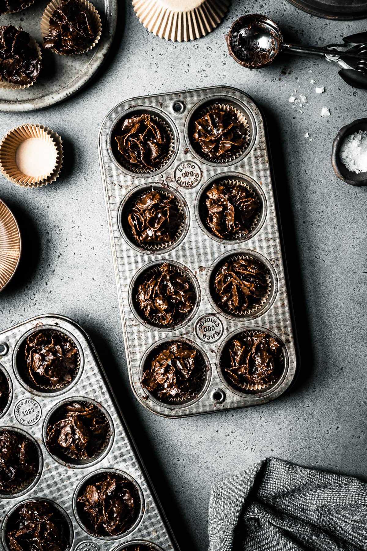 Two vintage mini muffin tins hold chocolate covered cornflake cereal, called roses de sables. They rest on a grey stone surface and are surrounded by brown bowls, brown paper muffin liners, a cookie scoop and a pinch bowl of flaky sea salt.