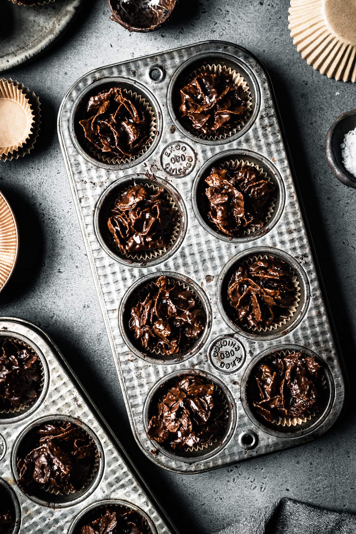 Two vintage mini muffin tins hold chocolate covered cornflake cereal, called roses de sables. They rest on a grey stone surface and are surrounded by brown bowls, brown paper muffin liners and a pinch bowl of flaky sea salt.
