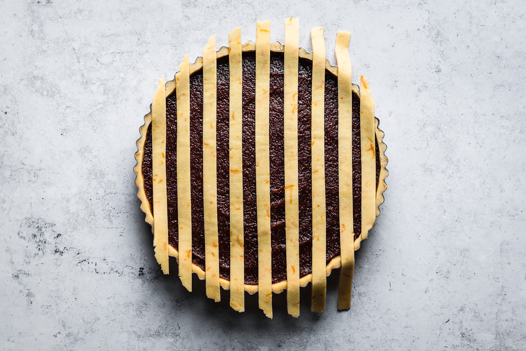 A process photo showing thin strips of lattice dough arranged vertically on a quince filled tart. The tart is on a blue grey surface.