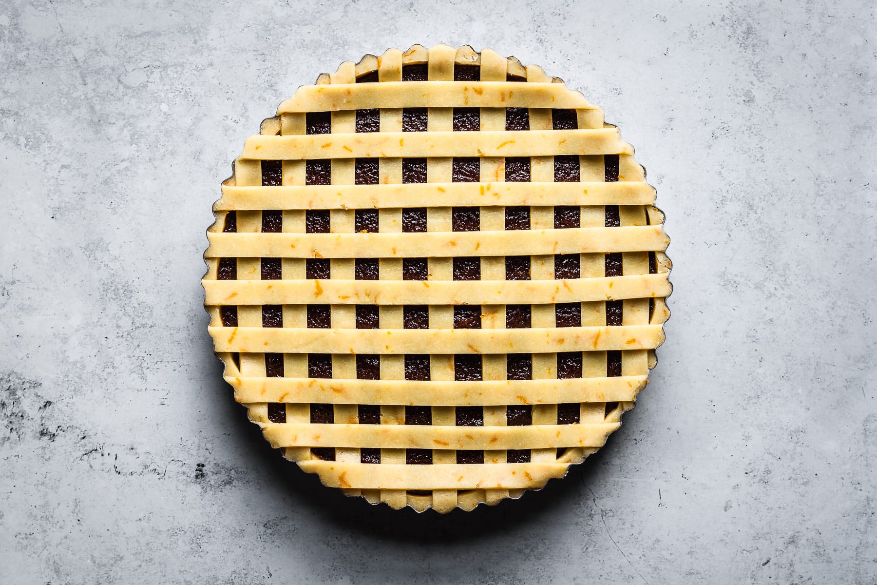 A process photo showing thin strips of lattice dough arranged horizontally (with vertical strips underneath) on a quince filled tart. The tart is on a blue grey surface.