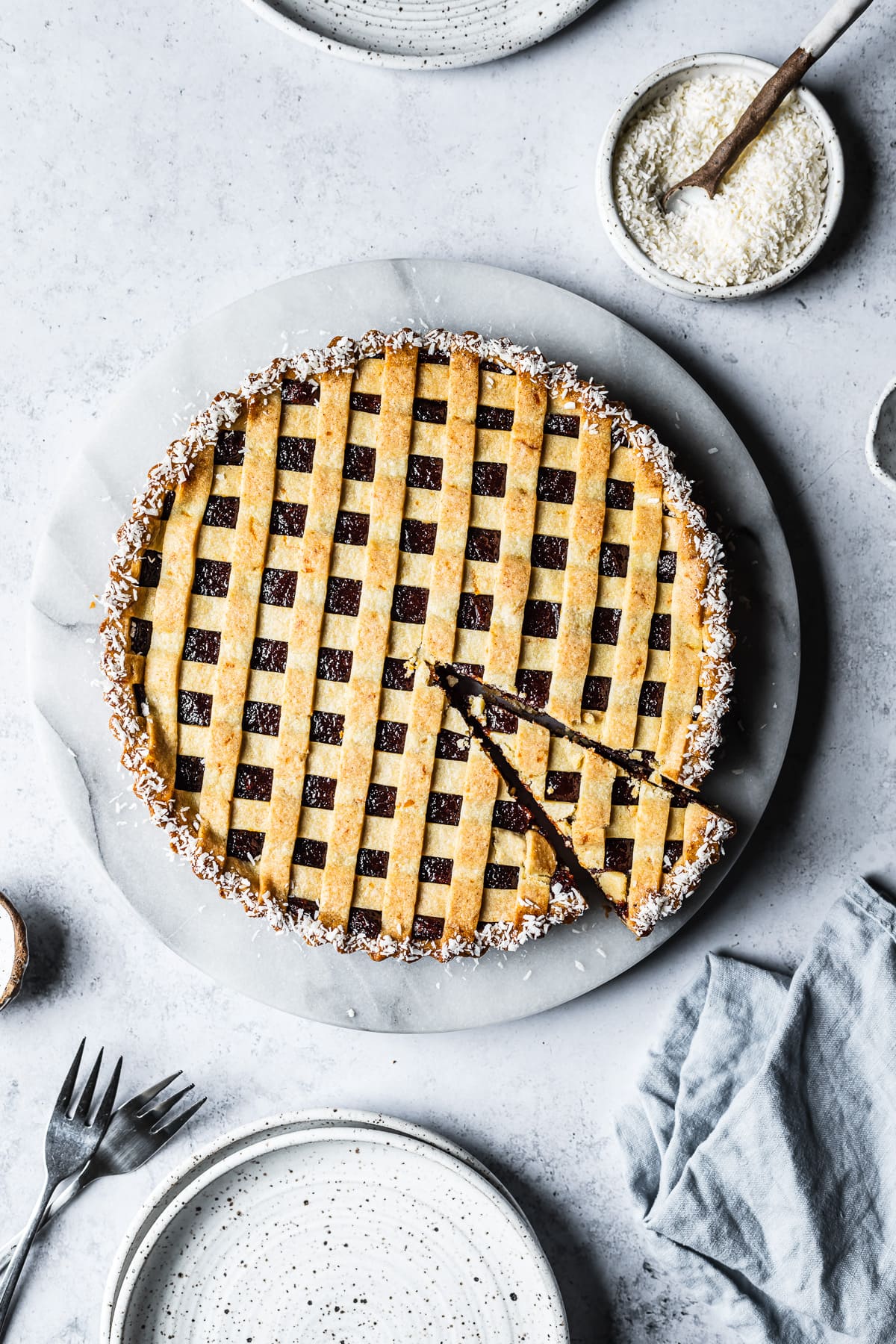 A baked pasta frola (quince tart) with thin lattice on top and a border of dessicated coconut. There is one slice cut from the tart. The tart rests on a marble platter on a blue-grey stone surface and is surrounded by small bowls, ceramic plates and a blue linen napkin.