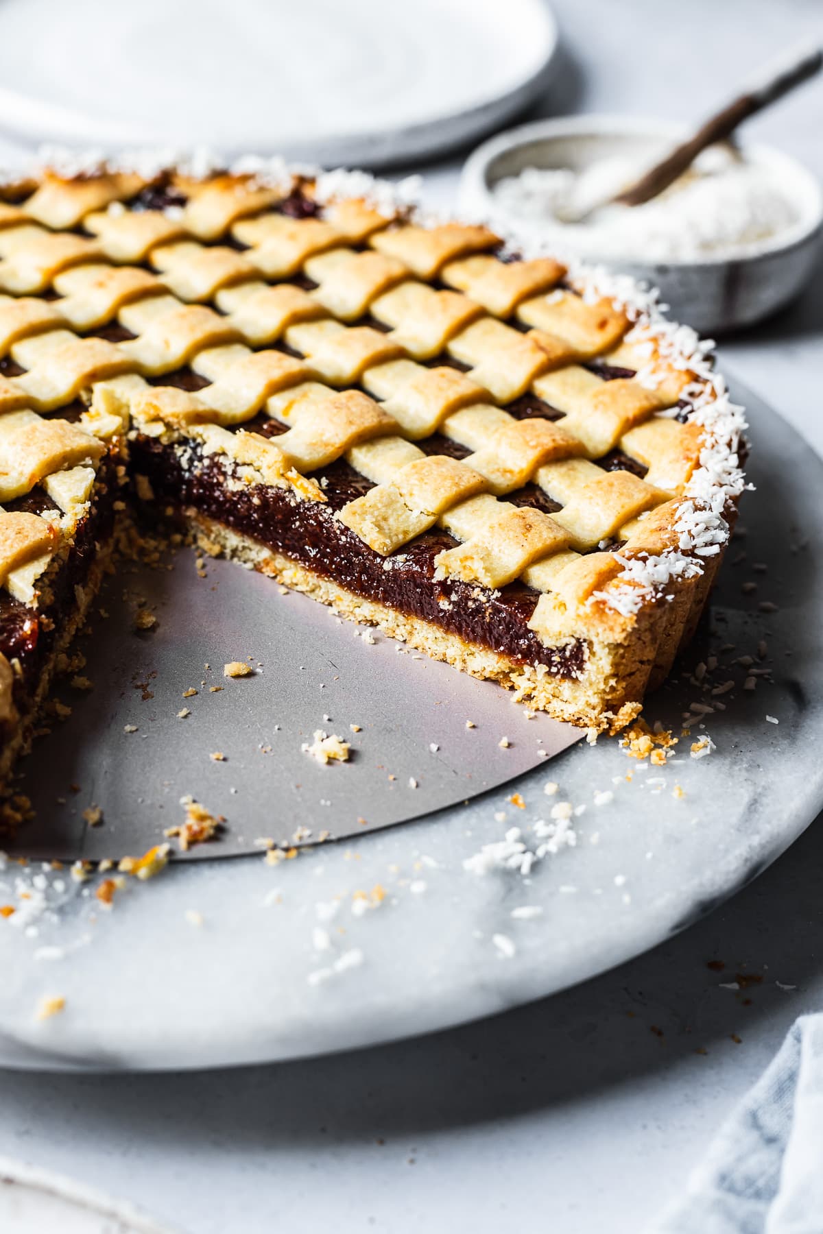 An angled view of a quince tart with lattice top and border of dessicated coconut. There are a few slices missing, revealing the base crust and filling. The tart rests on a marble platter on a blue grey surface. There is a bowl of coconut in the background next to a white ceramic plate.