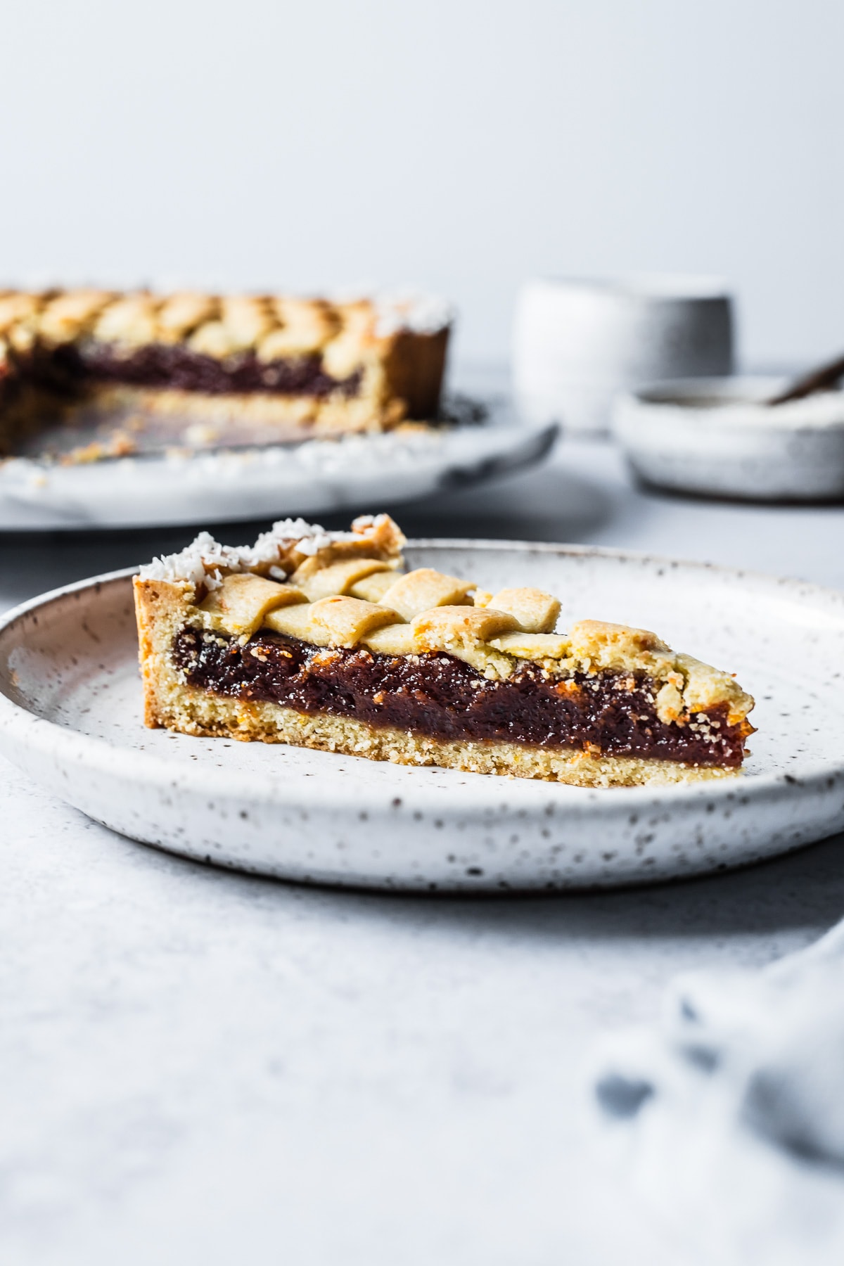 A front view of a slice of quince tart on a speckled white ceramic plate. The rest of the tart is out of focus in the background on a marble platter. There are ceramic mugs and bowls at back right. The plate rests on a blue grey stone surface.