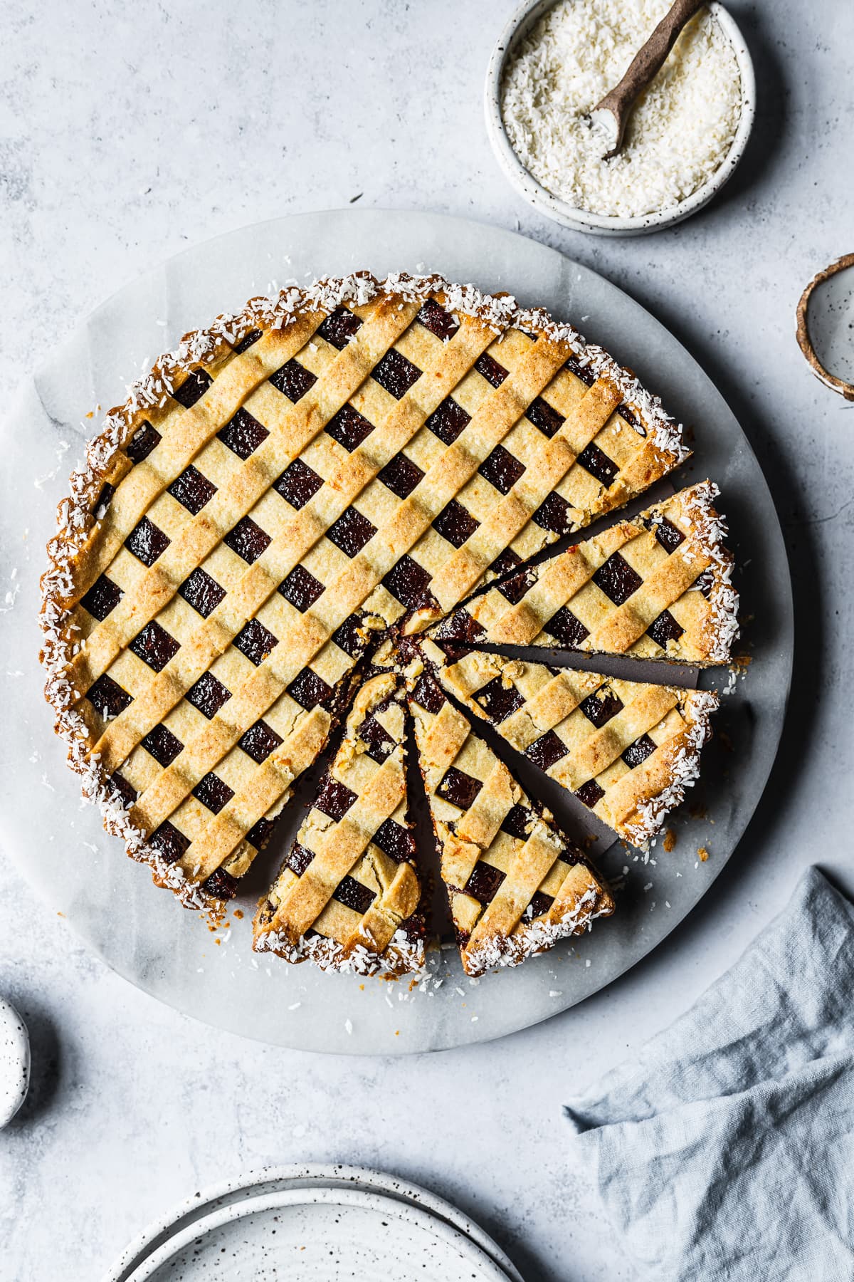 A baked pasta frola (quince tart) with thin lattice on top and a border of dessicated coconut. There are four slices cut from the tart. The tart rests on a marble platter on a blue-grey stone surface and is surrounded by small bowls, ceramic plates and a blue linen napkin.